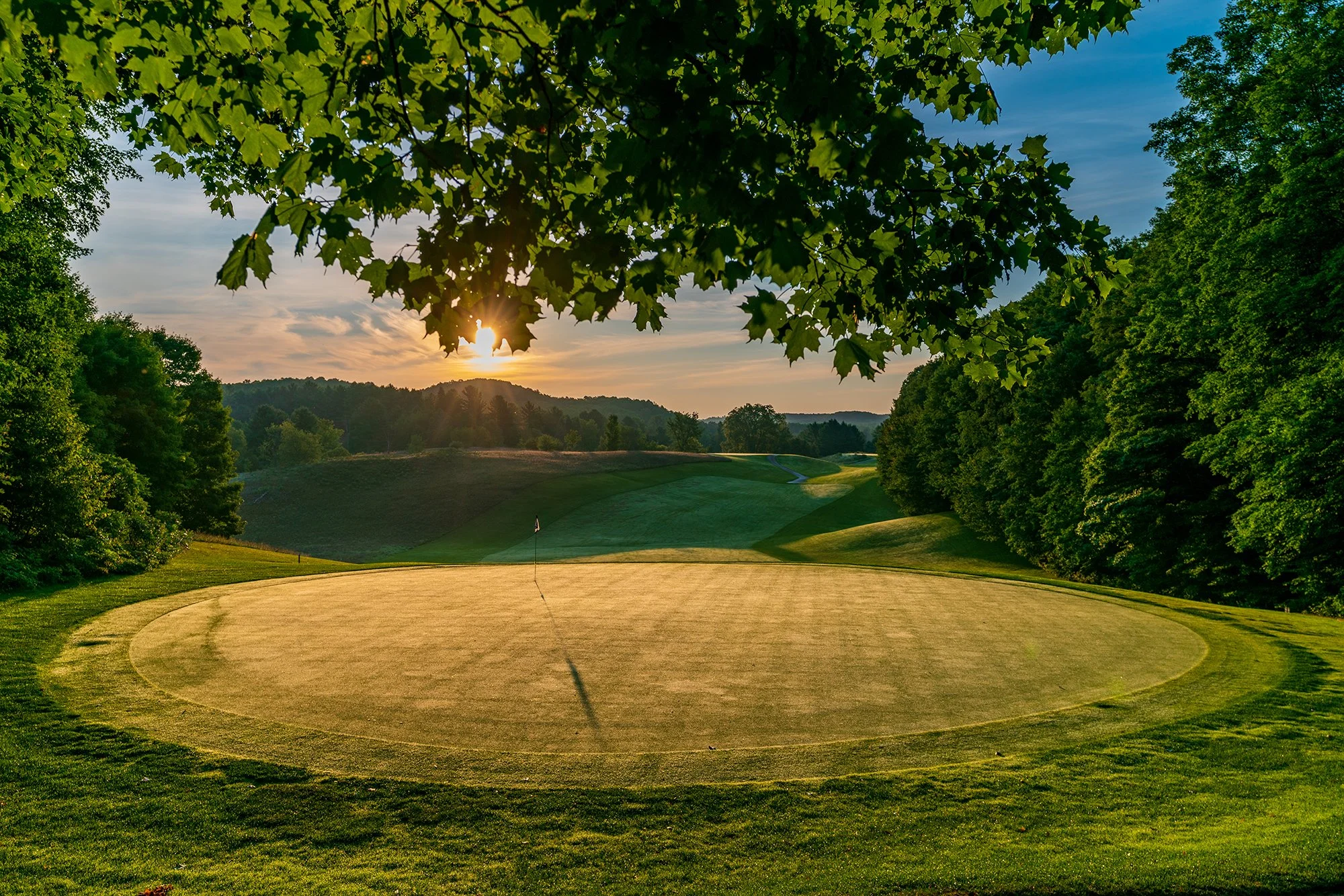 Sunset over a golf course with a green, flag, and surrounding trees.