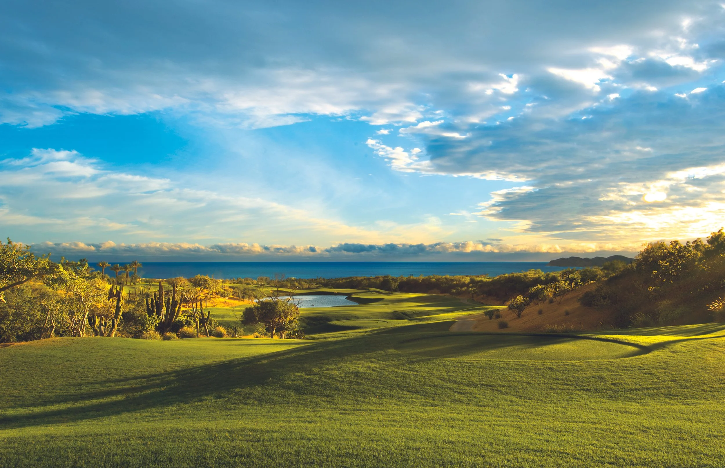 Scenic view of a golf course with lush green fairways, trees, and cacti, overlooking the ocean with a partly cloudy sky and distant mountains in the background.