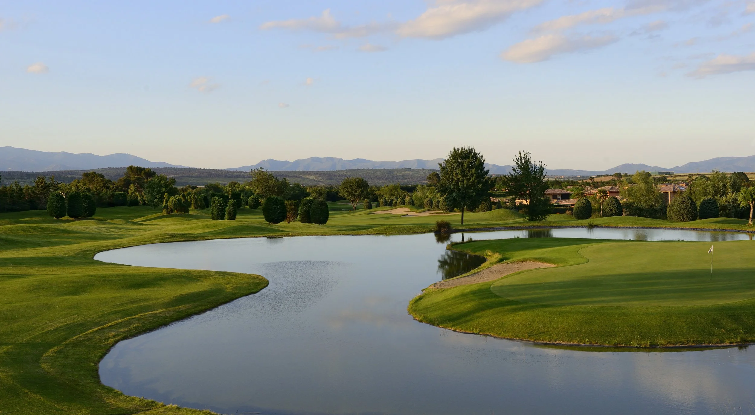 A golf course with green fairways, water hazards, trees, and distant mountains under a partly cloudy sky.