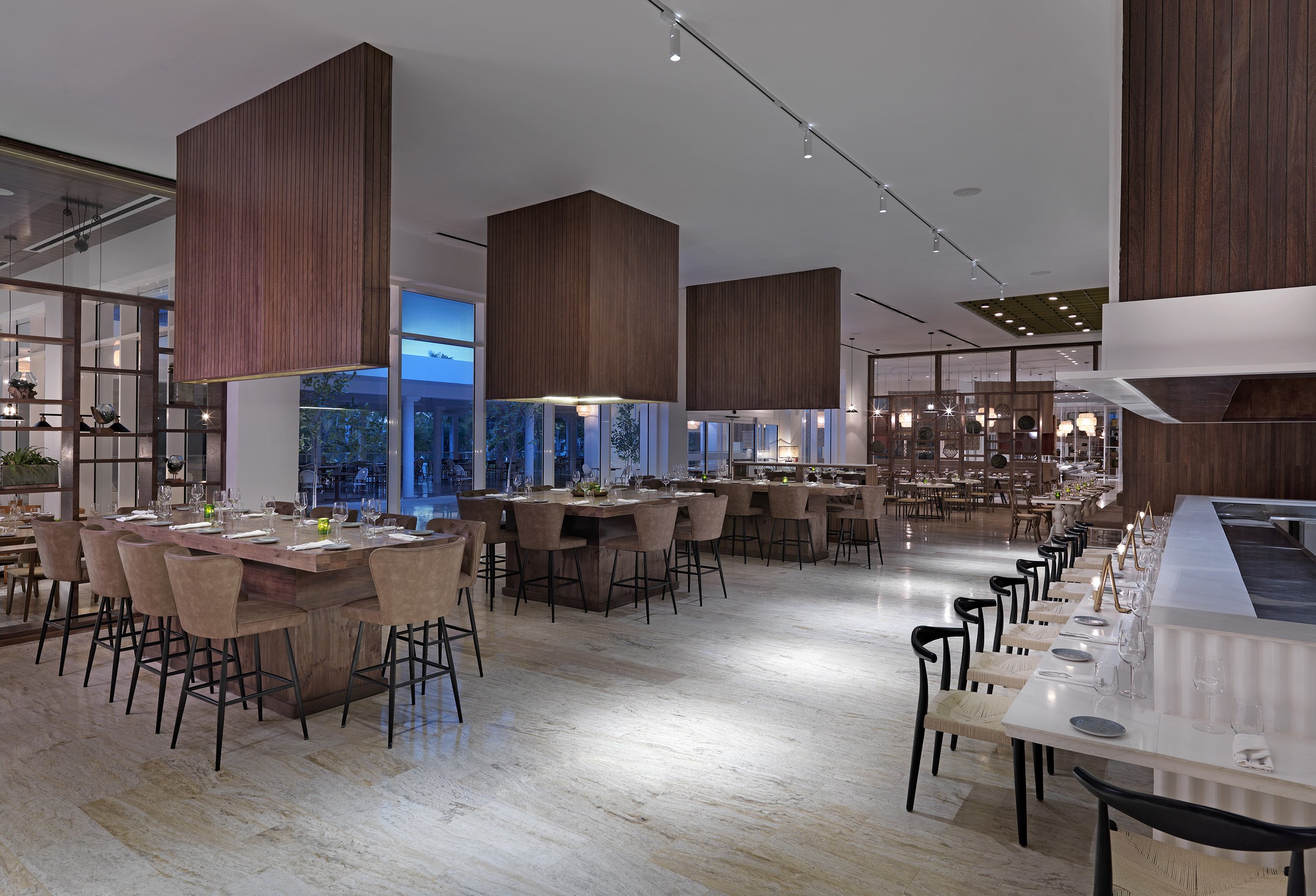 Elegant restaurant interior with wood-paneled columns, modern lighting, and neatly set tables with glassware and napkins, featuring large windows and a polished marble floor.