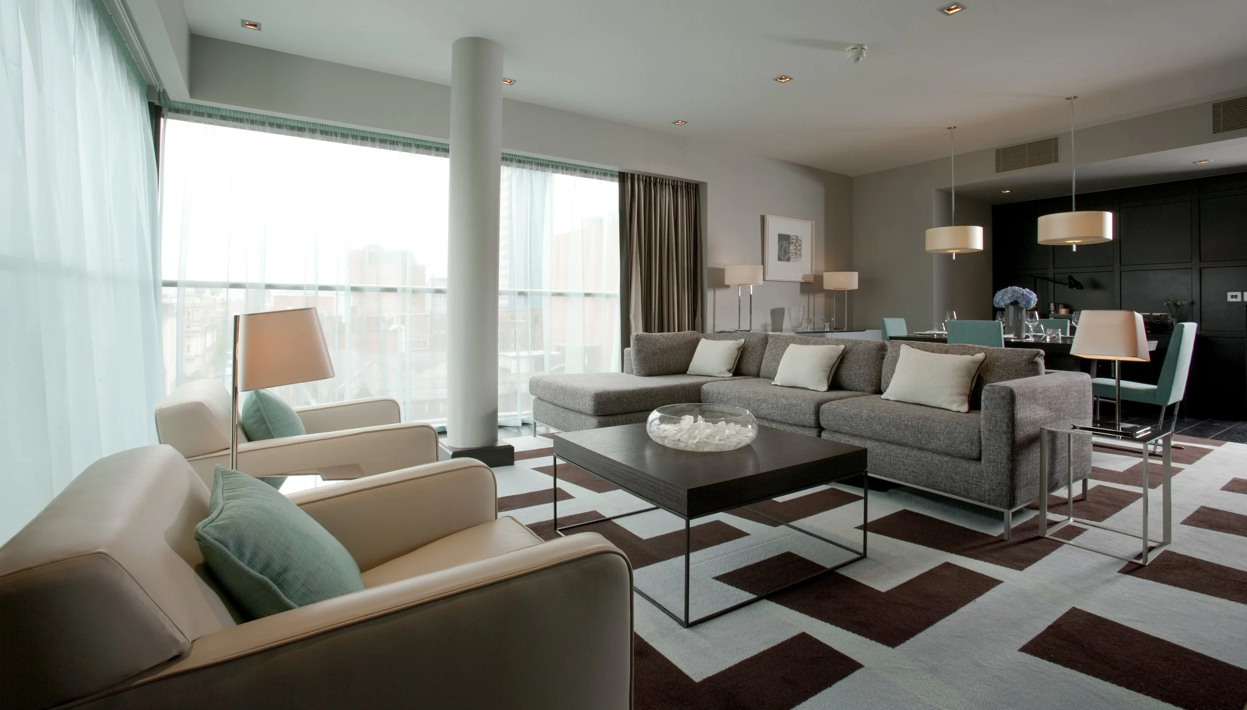 Modern living room with large windows, beige and gray furniture, a black coffee table, white pillows, a glass bowl, a patterned rug, and a dining area with teal chairs and pendant lights.