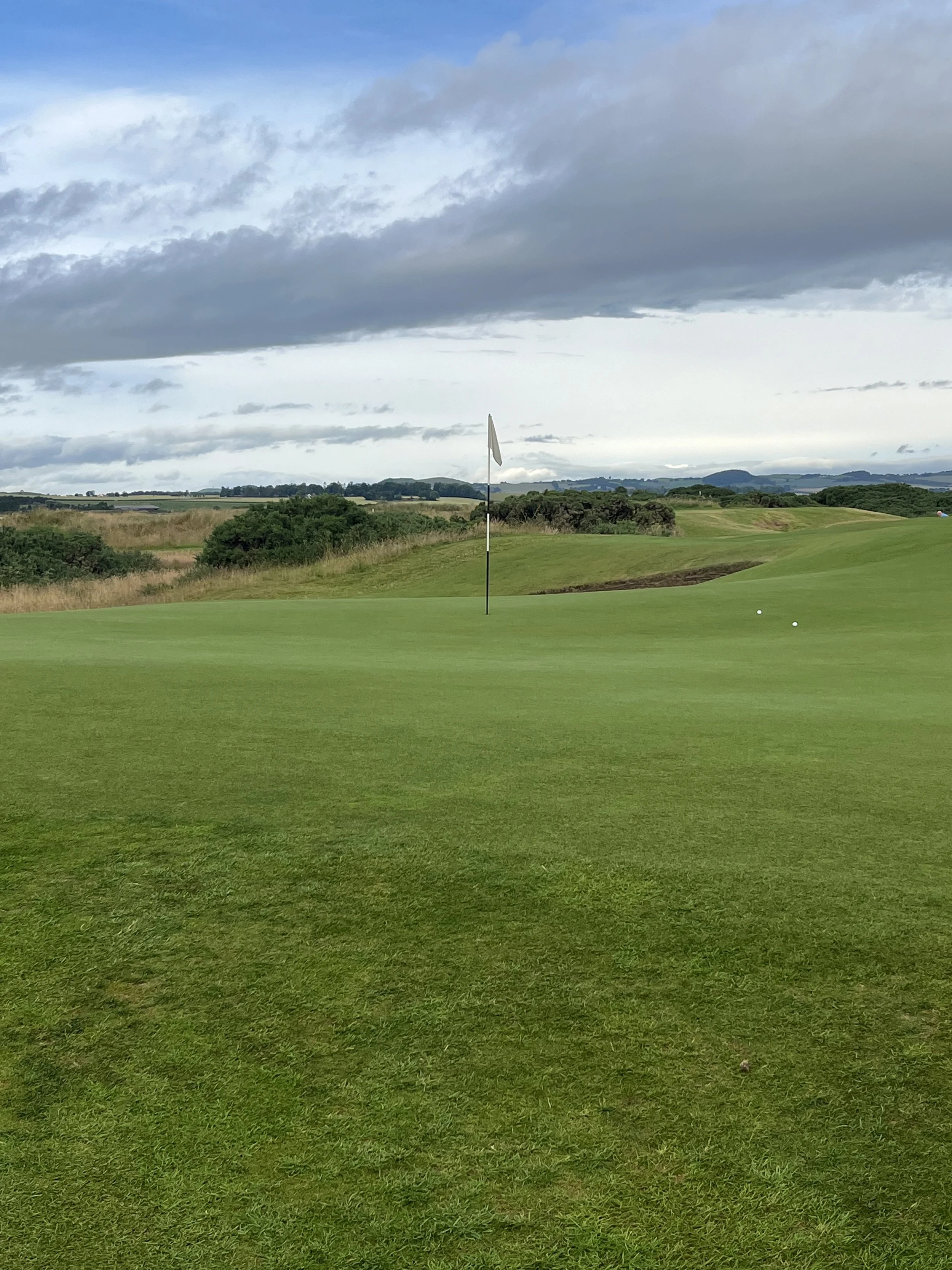 View of a golf course green with a golf flagstick and two golf balls, surrounded by rolling hills and cloudy sky.