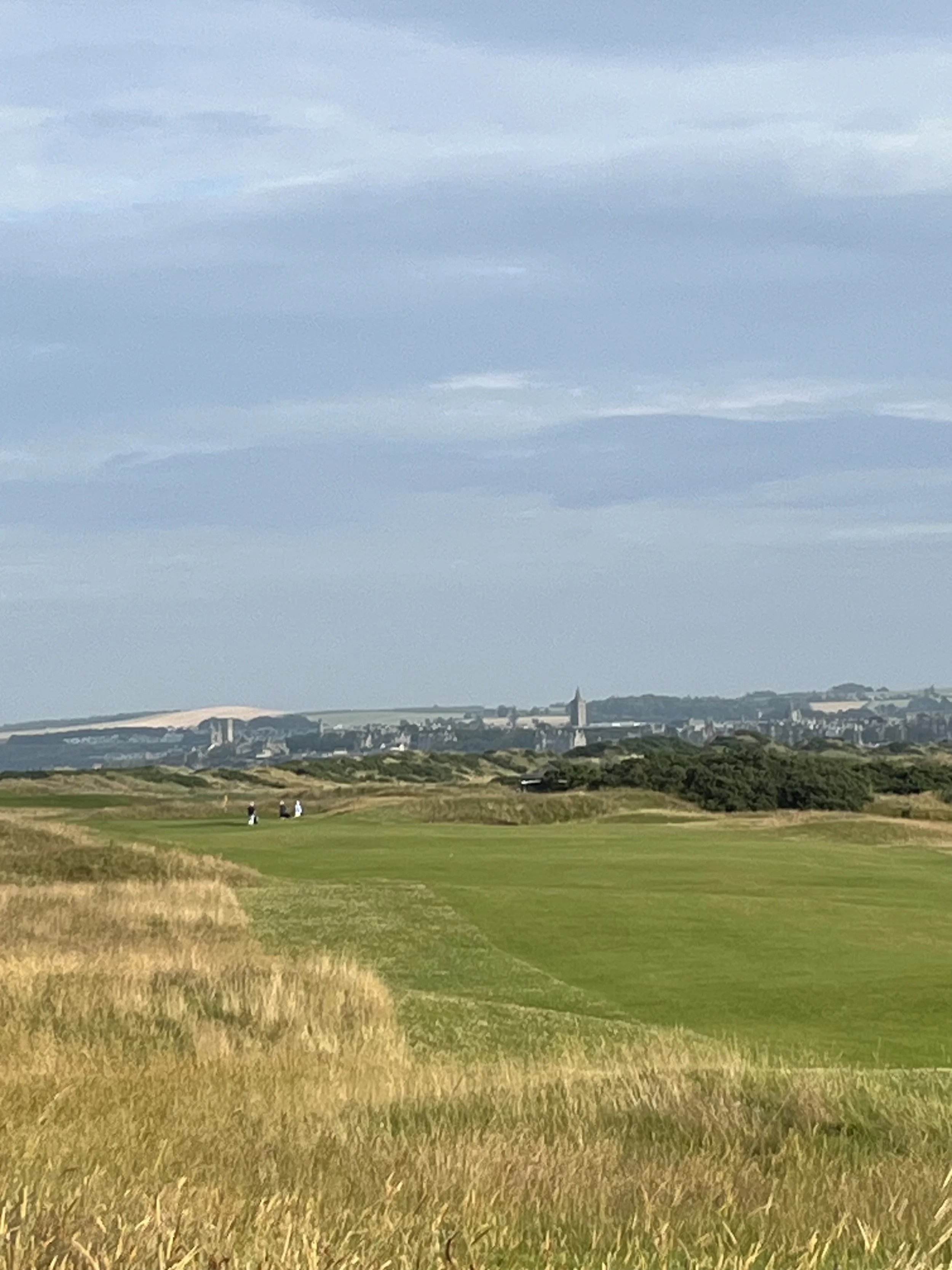 A scenic view of a golf course with a cityscape in the background, under a partly cloudy sky.