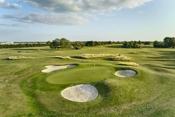 Golf course landscape with green, sand bunkers, and trees under a partly cloudy sky.