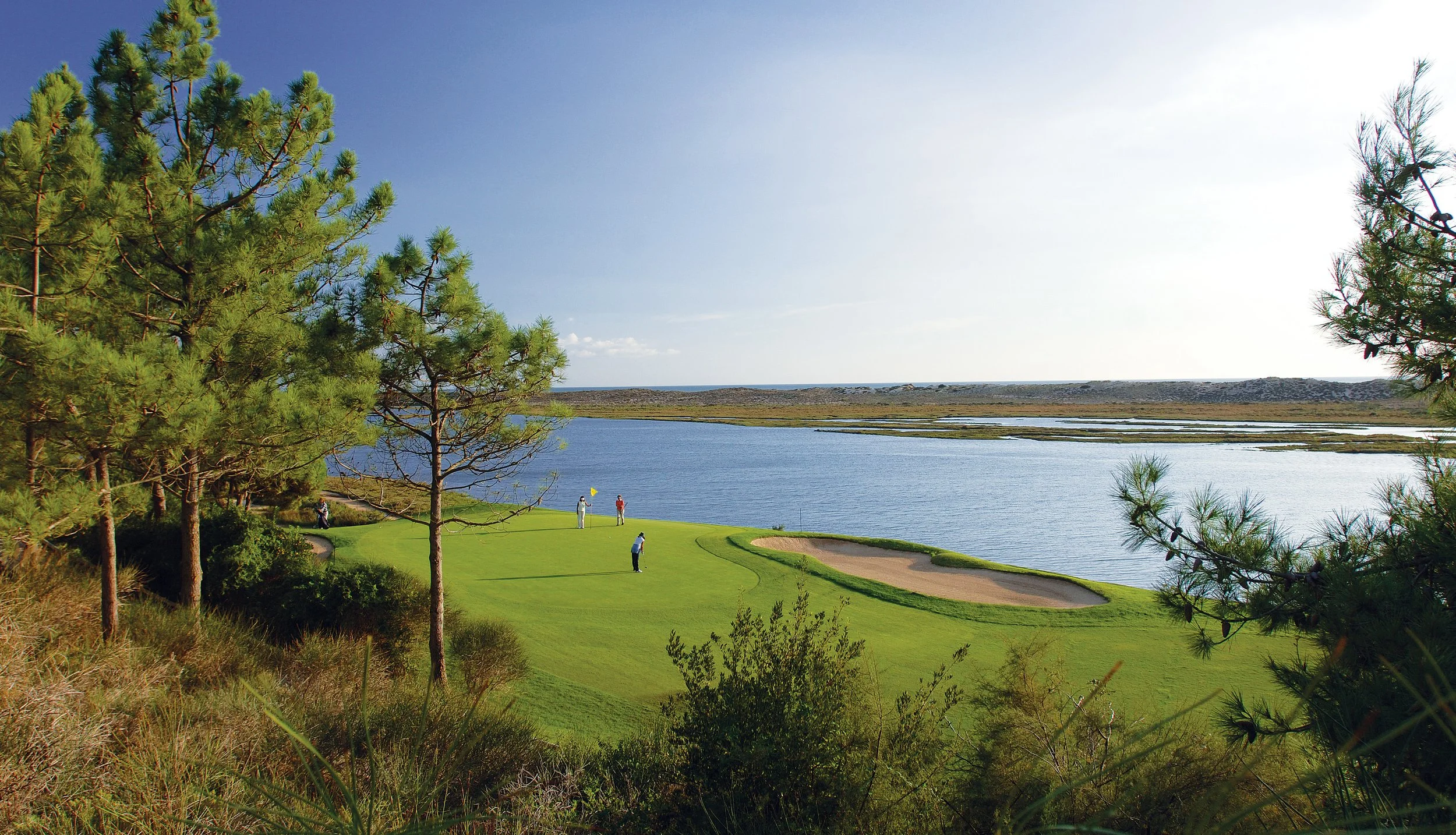 A scenic view of a golf course next to a large body of water, with trees in the foreground and three golfers on the green.