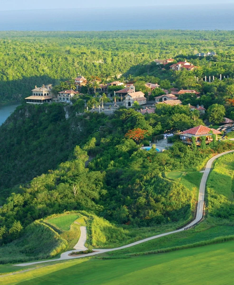 Aerial view of a lush, green landscape with buildings resembling a village on a hilltop, surrounded by dense forest and overlooking the ocean in the distance.