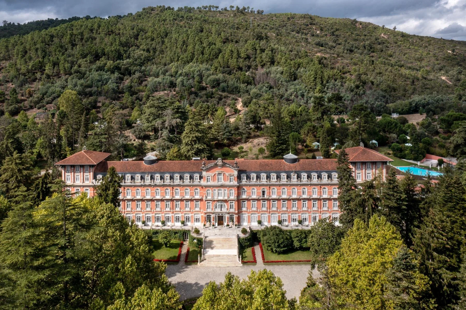 A large, ornate pink and white mansion with many windows, set amidst lush greenery, with a hillside covered in trees and shrubs in the background.