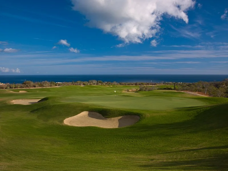 Scenic view of a golf course with green fairways, sand bunkers, and the ocean in the background under a partly cloudy sky.