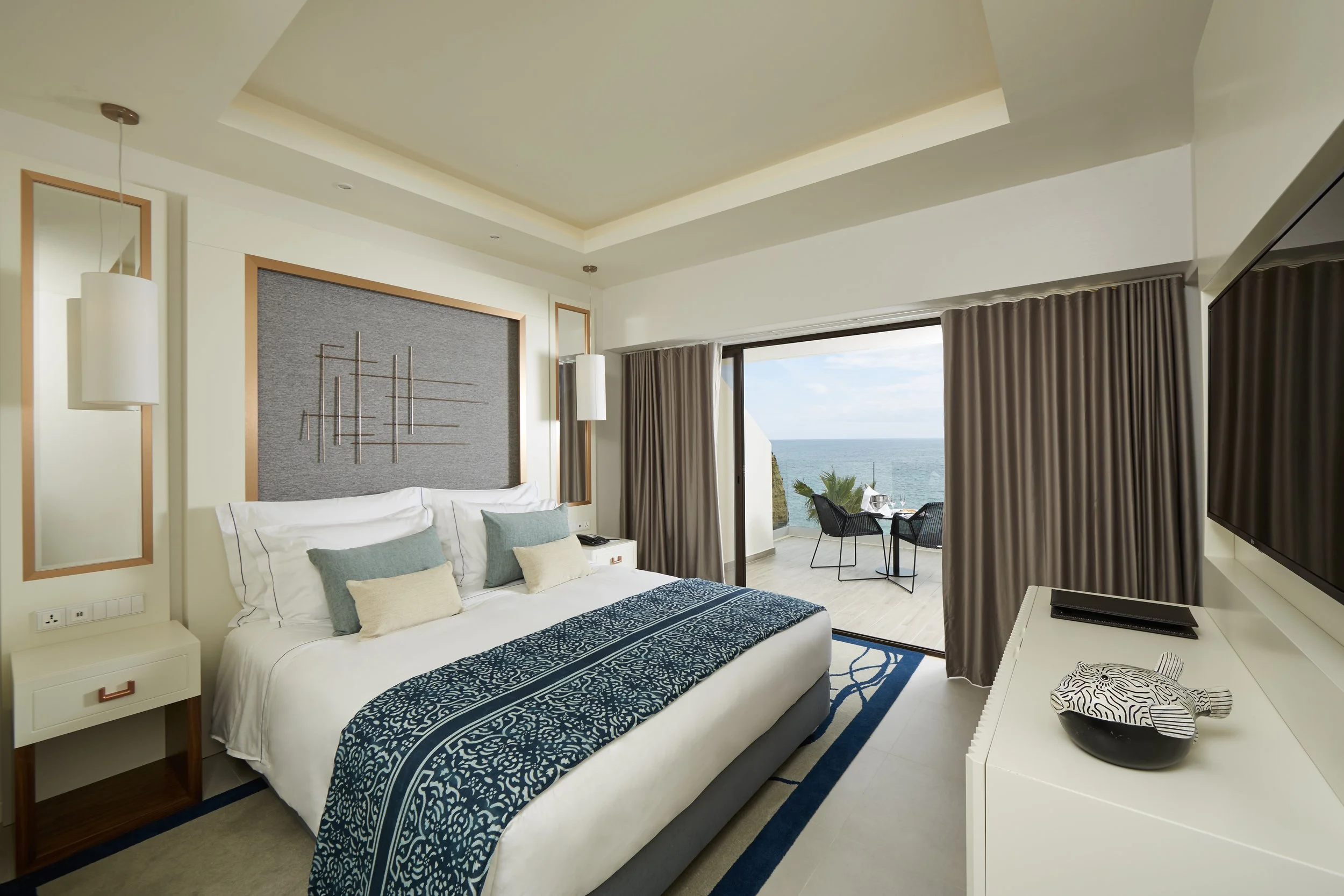 Bedroom with a view of the ocean, containing a large bed with pillows and a decorative bed runner, a balcony with a small table and chairs, and a television.