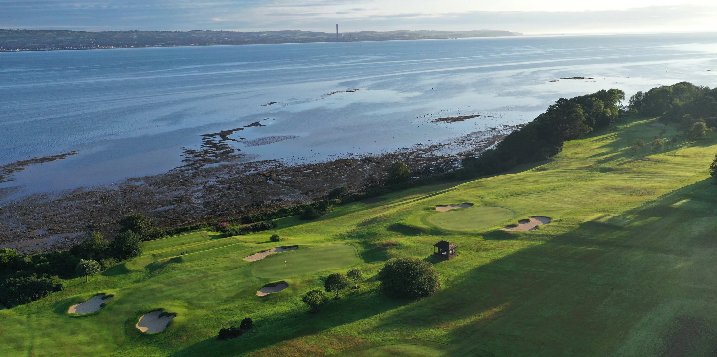 Aerial view of a golf course situated by a coastline with water and rocky shores; trees and fairways are visible.
