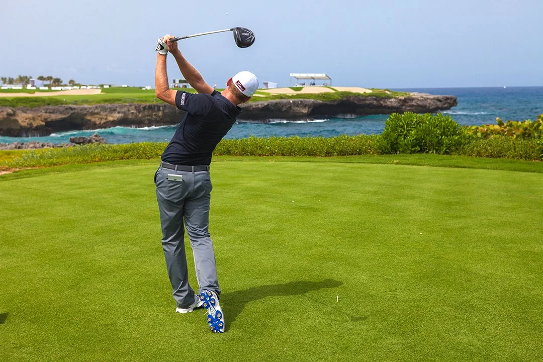 A man playing golf on a lush green course near the ocean, swinging a golf club.