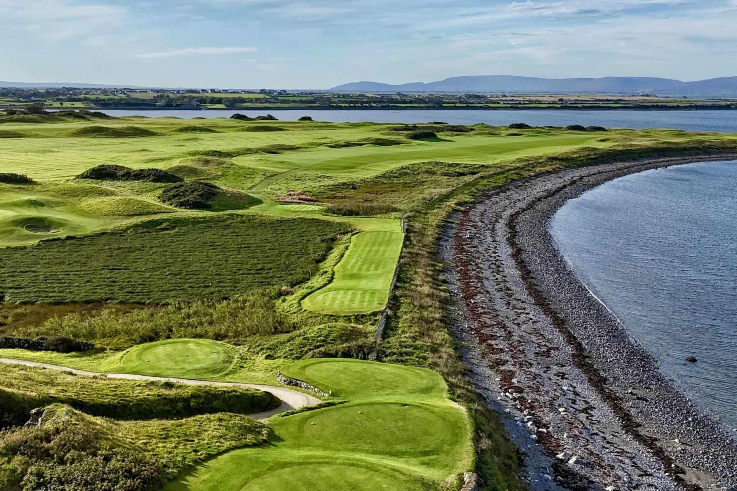 "An aerial view of The Hawthorn at Galway Bay, featuring vibrant green golf course tee boxes and fairways curving along a rocky shoreline, with calm blue waters and distant hills under a partly cloudy sky."