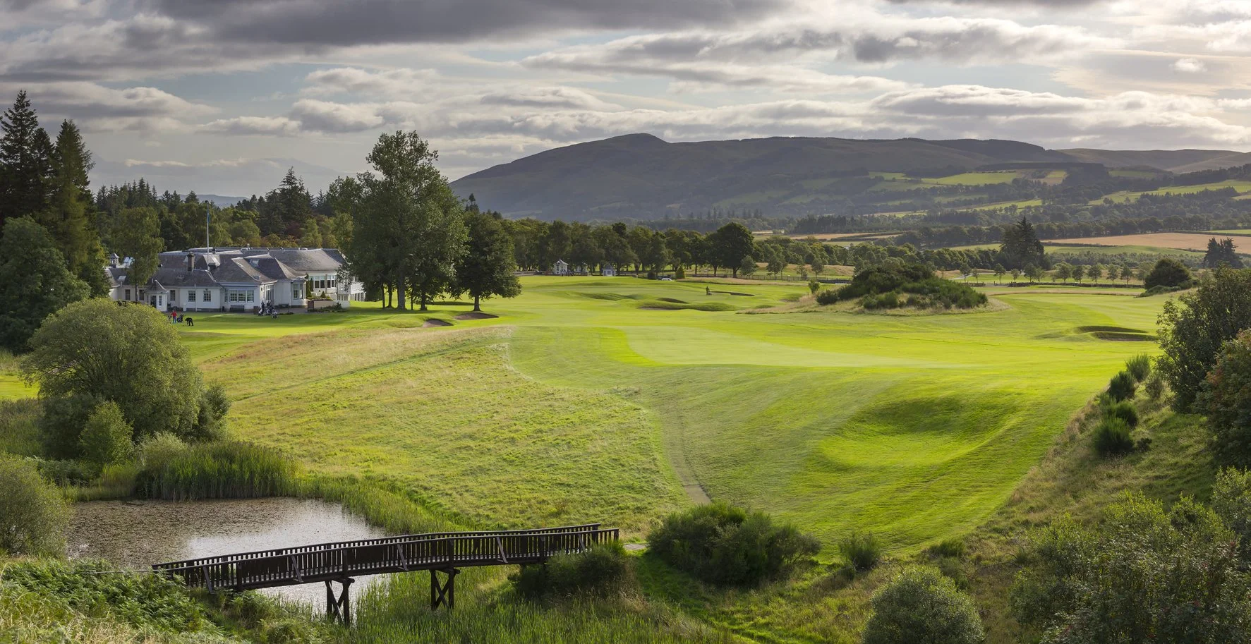 Scenic view of a golf course with a clubhouse, lush green fairways, trees, a small pond, and wooden bridge under a partly cloudy sky.