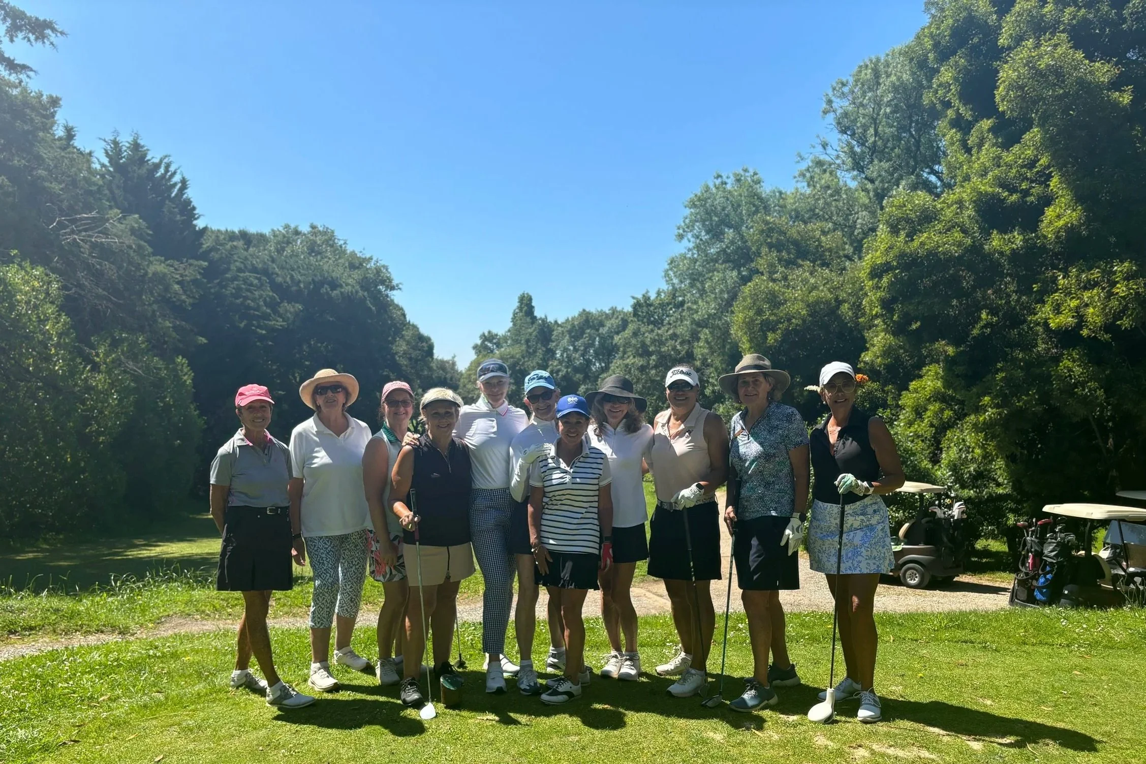 Group of women standing on a golf course, holding golf clubs, wearing hats, sunglasses, and golf attire, with golf carts and trees in the background on a sunny day.