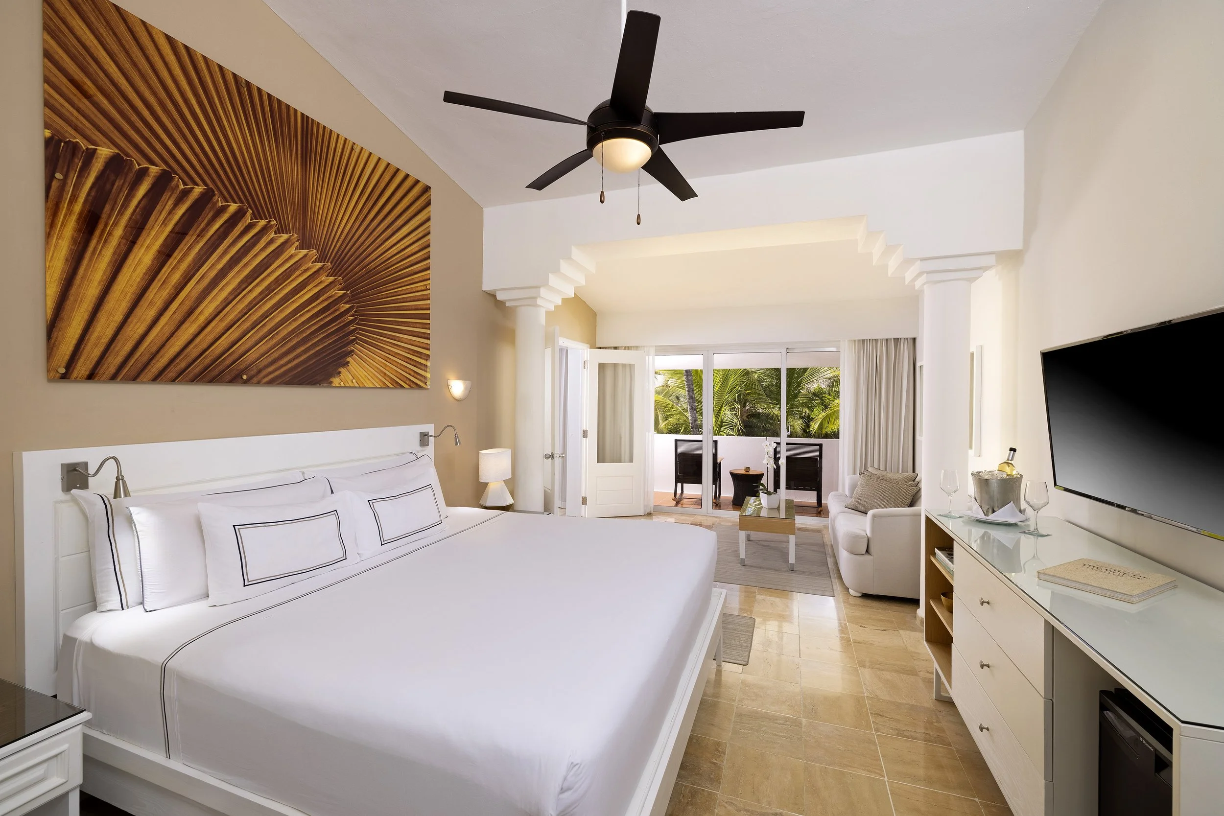 A spacious hotel room with a large white bed, a ceiling fan, a flat-screen TV, a sitting area with an armchair and a small table, and a balcony with outdoor seating and tropical plants.