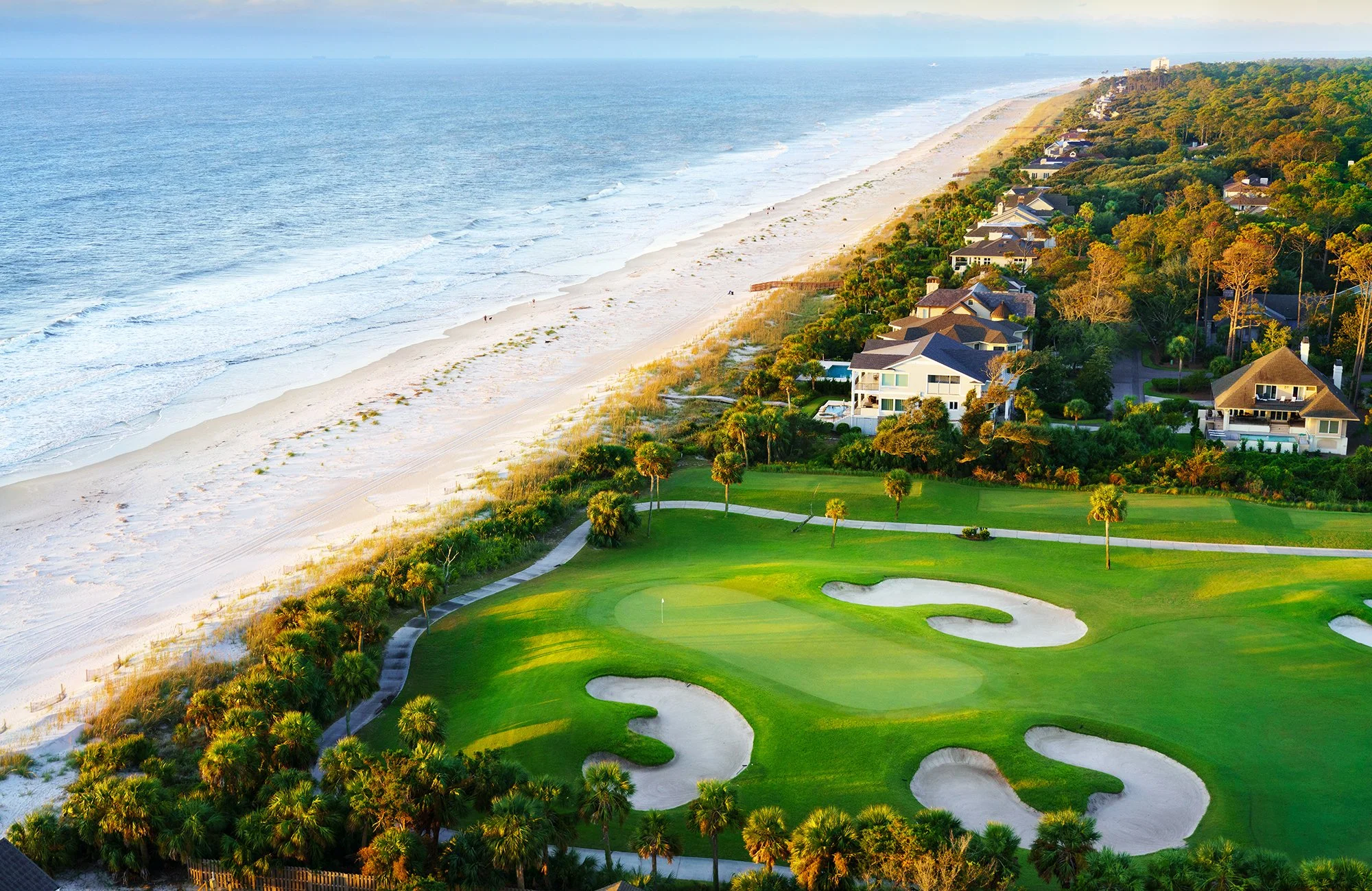Aerial view of a beachside golf course with sand traps, lush green grass, and palm trees, adjacent to a residential neighborhood along the coast with the ocean in the background.