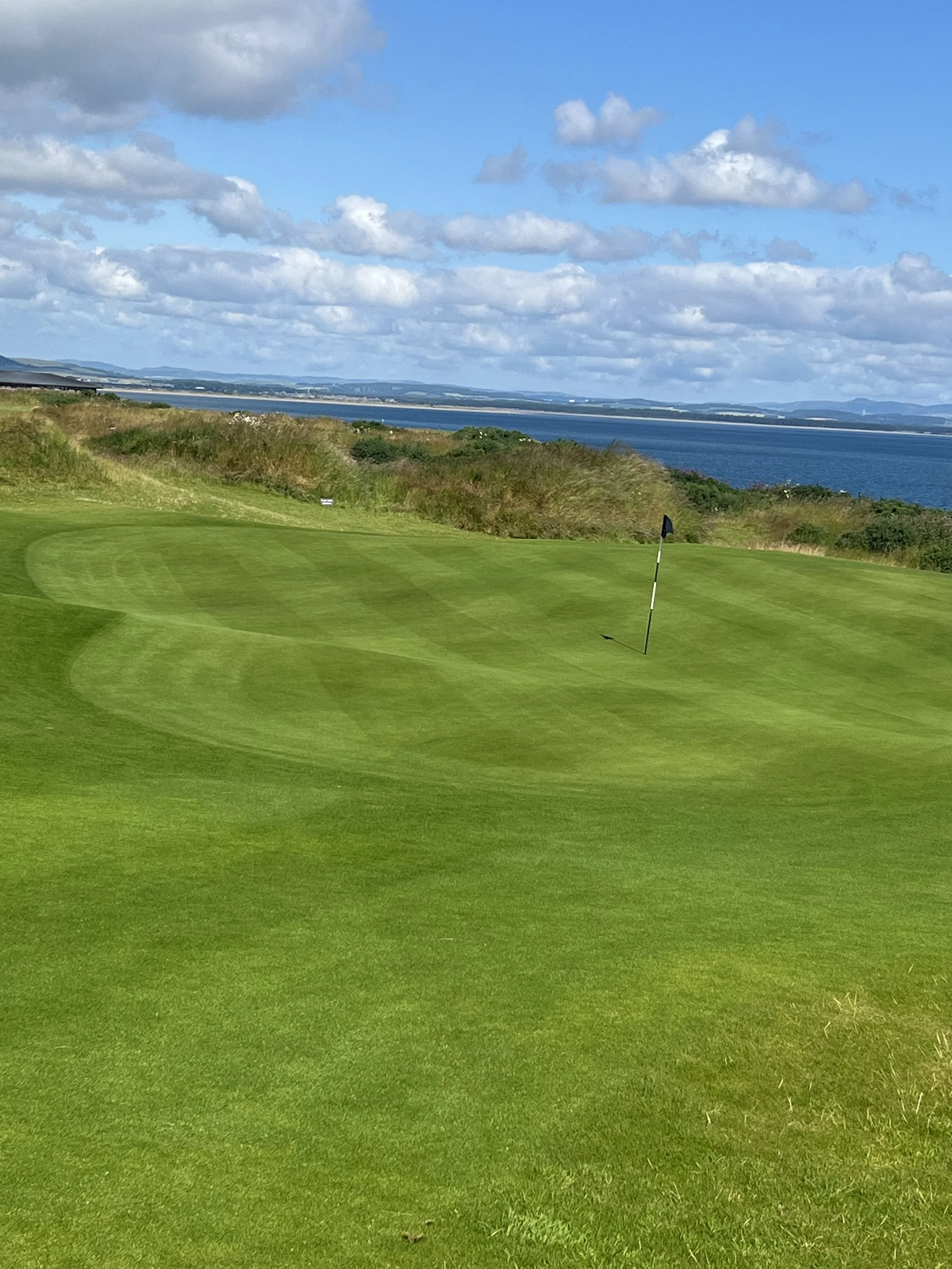 A view of a golf course green with a flagstick, surrounded by well-maintained grass, overlooking a body of water with hills in the distance, under a partly cloudy sky.