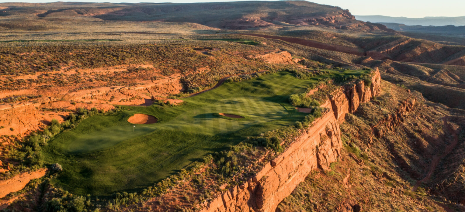 A golf course on a green hilltop surrounded by rocky red desert terrain and sparse vegetation.