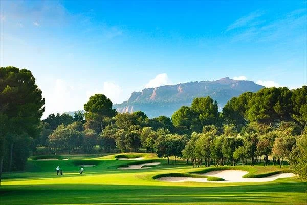 Golf course with green fairways, sand traps, and trees, with mountains in the background under a blue sky.