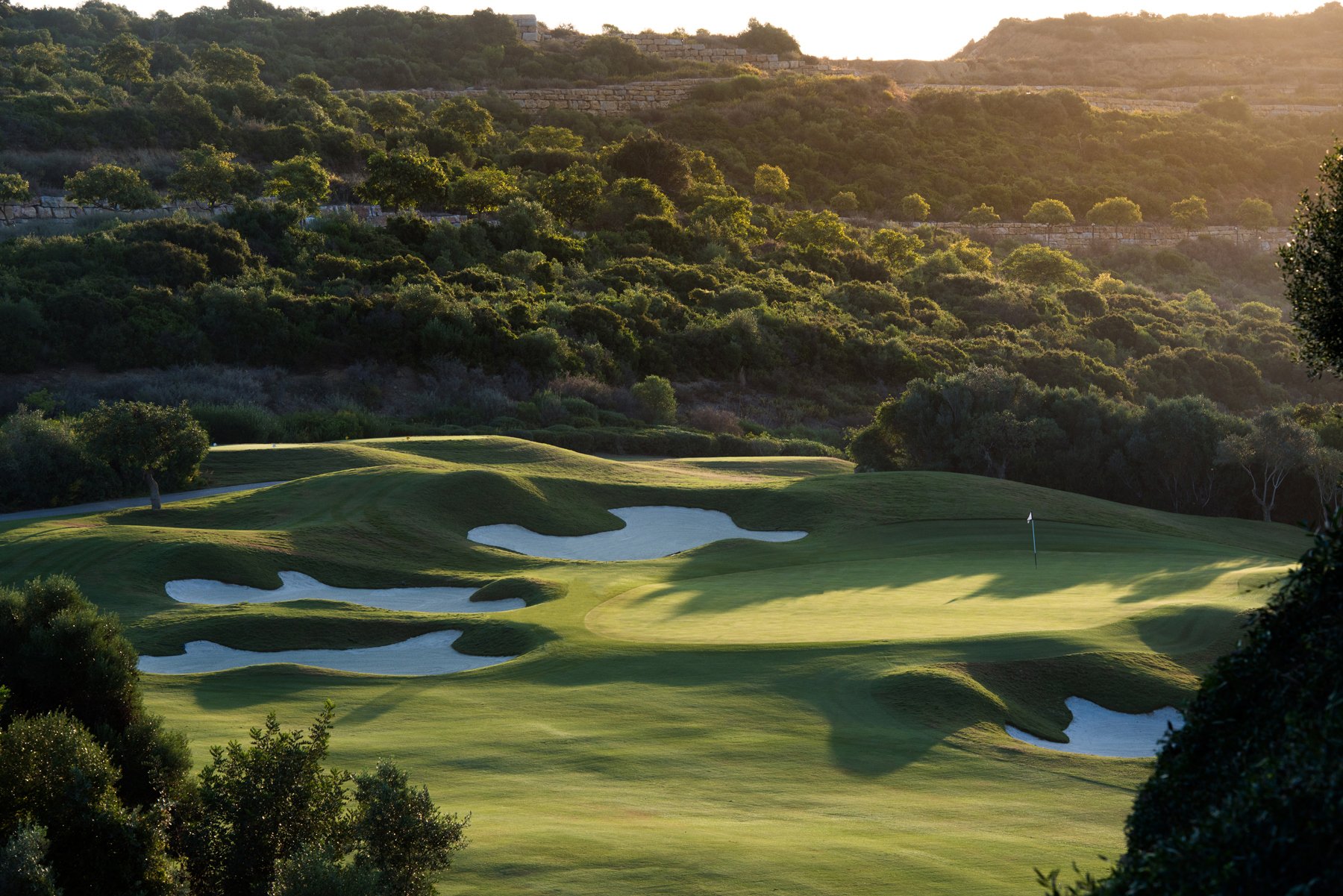 A golf course with green fairways and sand bunkers, surrounded by trees and hills, with a scenic landscape in the background at sunset.