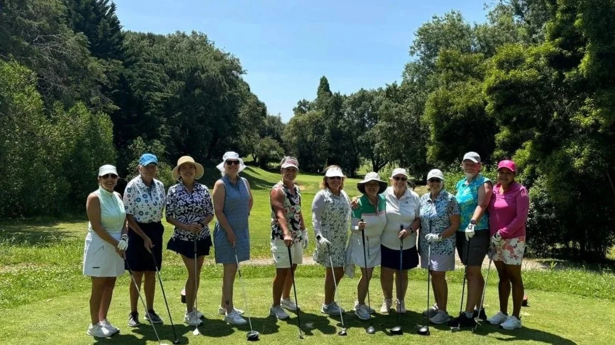 Group of women standing on a golf course holding golf clubs, dressed in sporty golf attire with hats and sunglasses, surrounded by trees under a clear sky.
