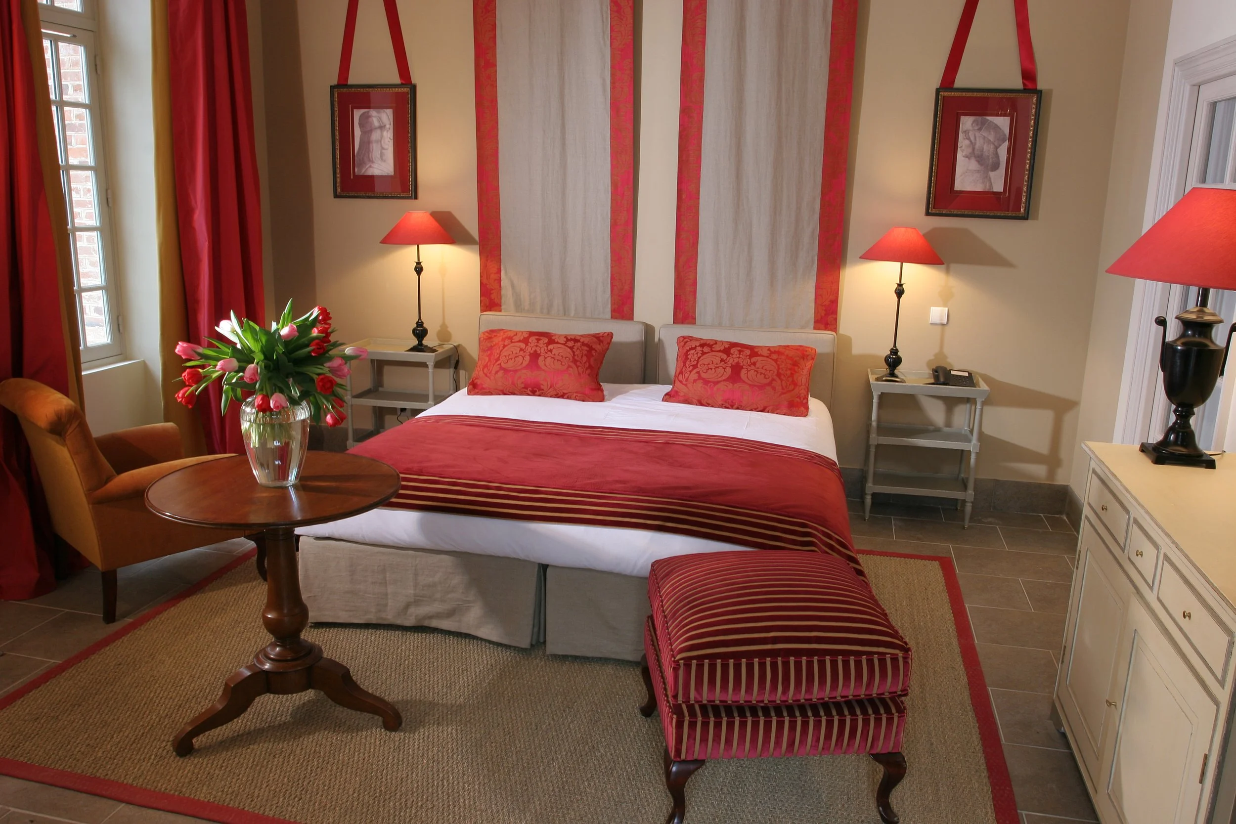 A hotel room with a double bed, red and beige decor, floral arrangements, and bedside lamps