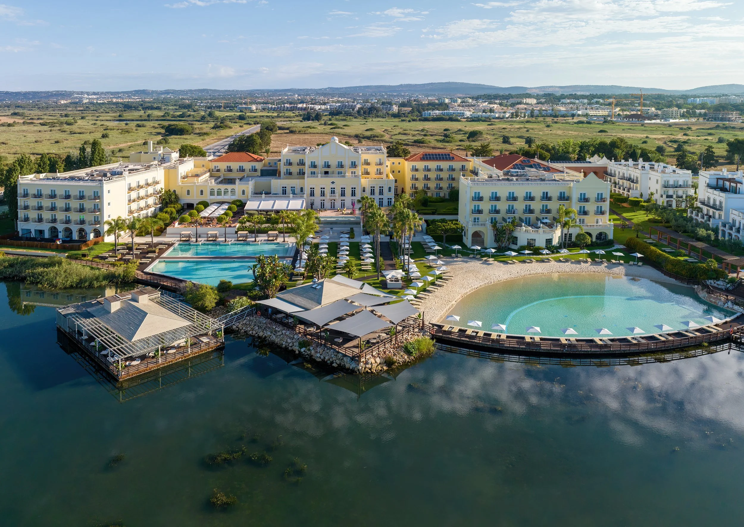 Aerial view of a luxurious resort with multiple pools, lounging areas, a water body, and white buildings with blue accents surrounded by greenery against a distant plain and hills.