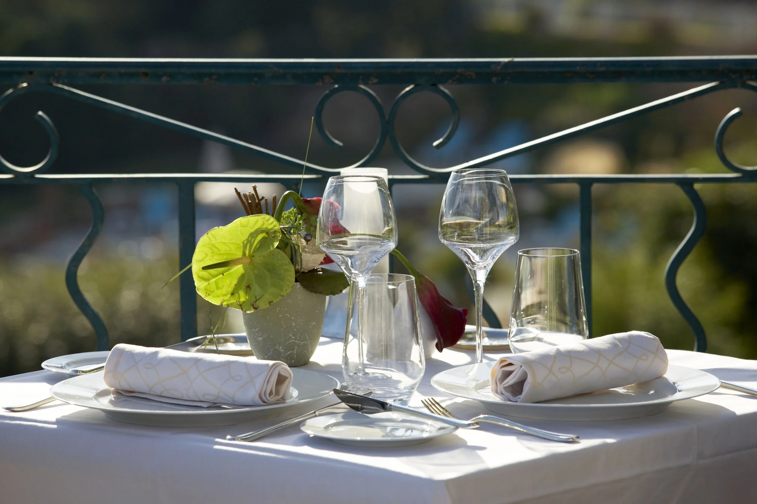 Elegant outdoor dining table set with white tablecloth, cloth napkins, wine glasses, and a white vase with green plants and pink flowers. The background shows a decorative metal railing and trees.