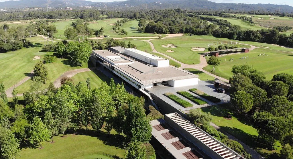 Aerial view of a modern building surrounded by a golf course with trees, pathways, and sand traps in a lush green landscape.