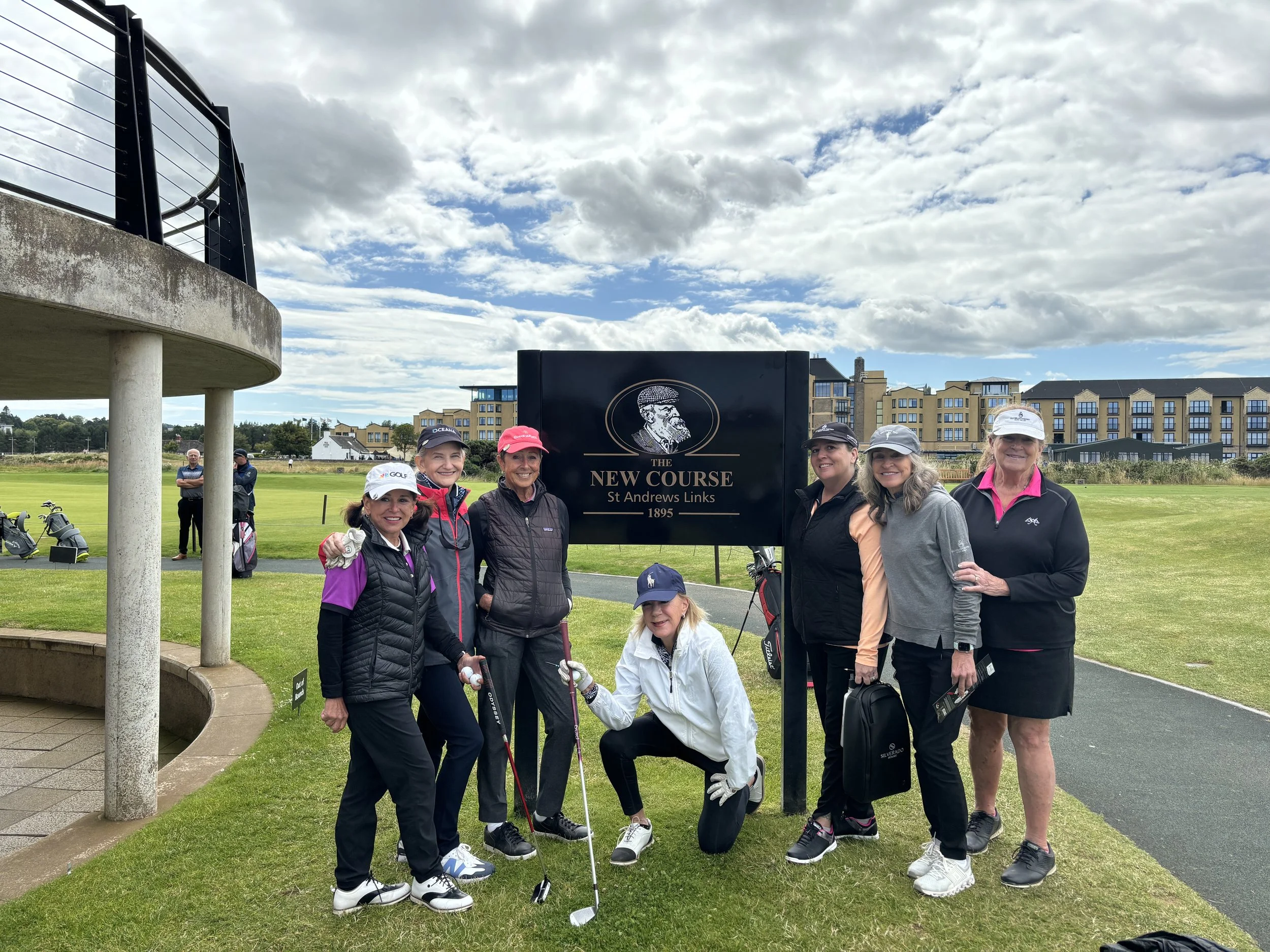 Group of golfers posing at St Andrews Links "New Course" sign, wearing golf attire, with buildings and cloudy sky in the background.