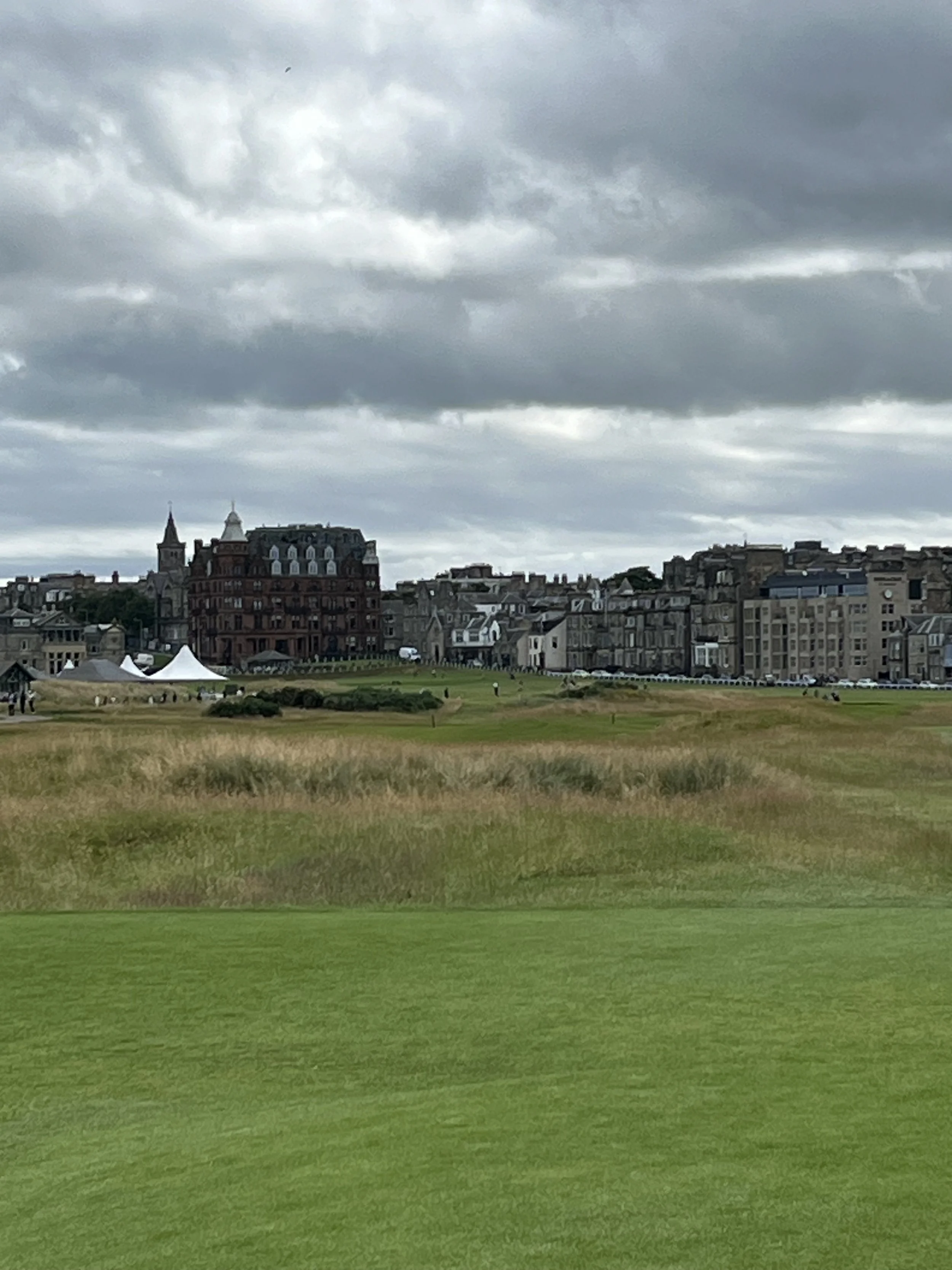 A view of a golf course with lush green grass in the foreground, brown and tall grass in the middle, and an old cityscape with historic buildings and tents in the background under a cloudy sky.