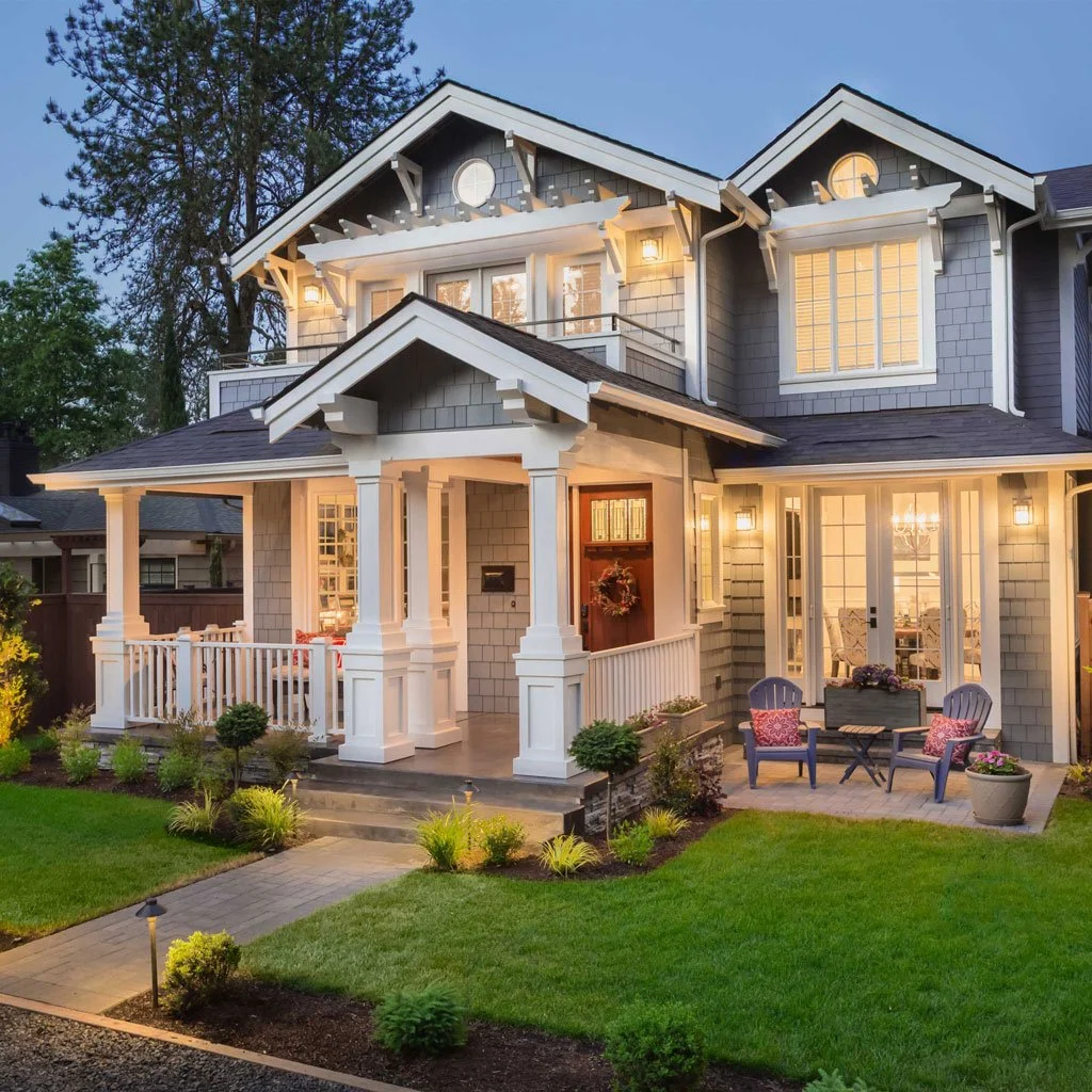 A two-story suburban house with gray siding, white trim, a covered front porch, illuminated windows, and a small outdoor seating area on a patio, surrounded by a well-manicured lawn and landscaped garden.