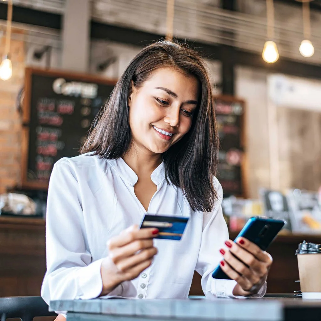 Young woman in a white blouse holding a credit card and a smartphone, smiling in a coffee shop with a chalkboard menu and warm lighting.