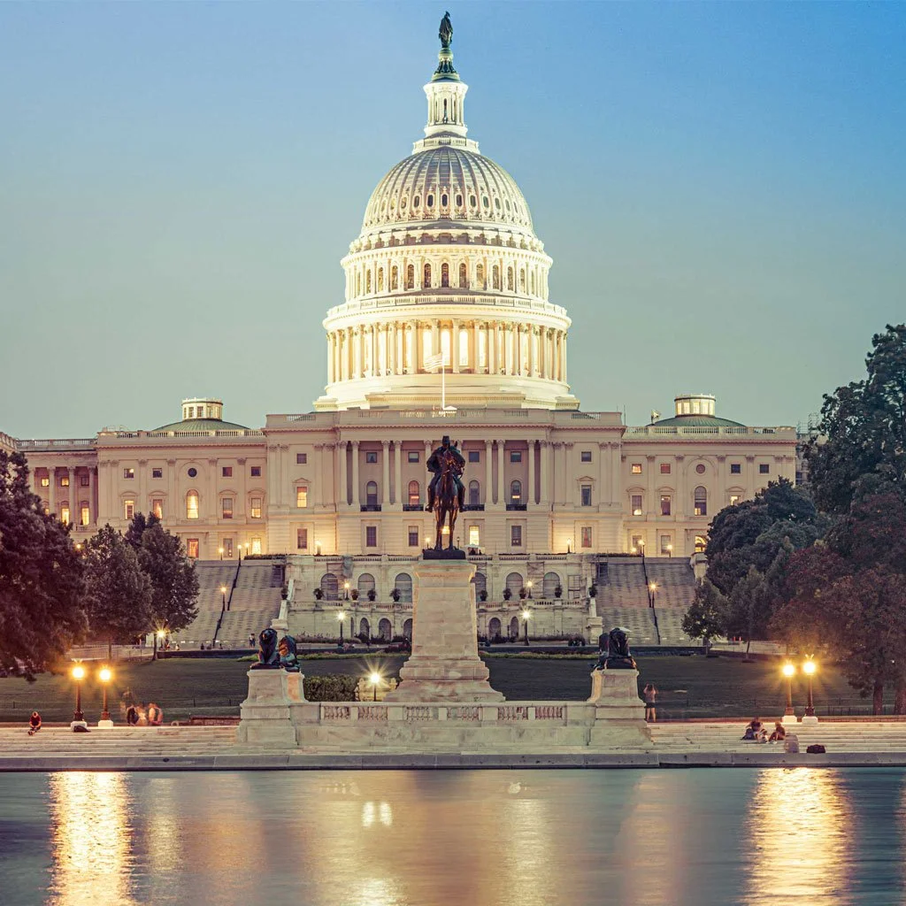 The United States Capitol building illuminated at dusk, with a pond and trees in the foreground, and a statue of a man on horseback in front.