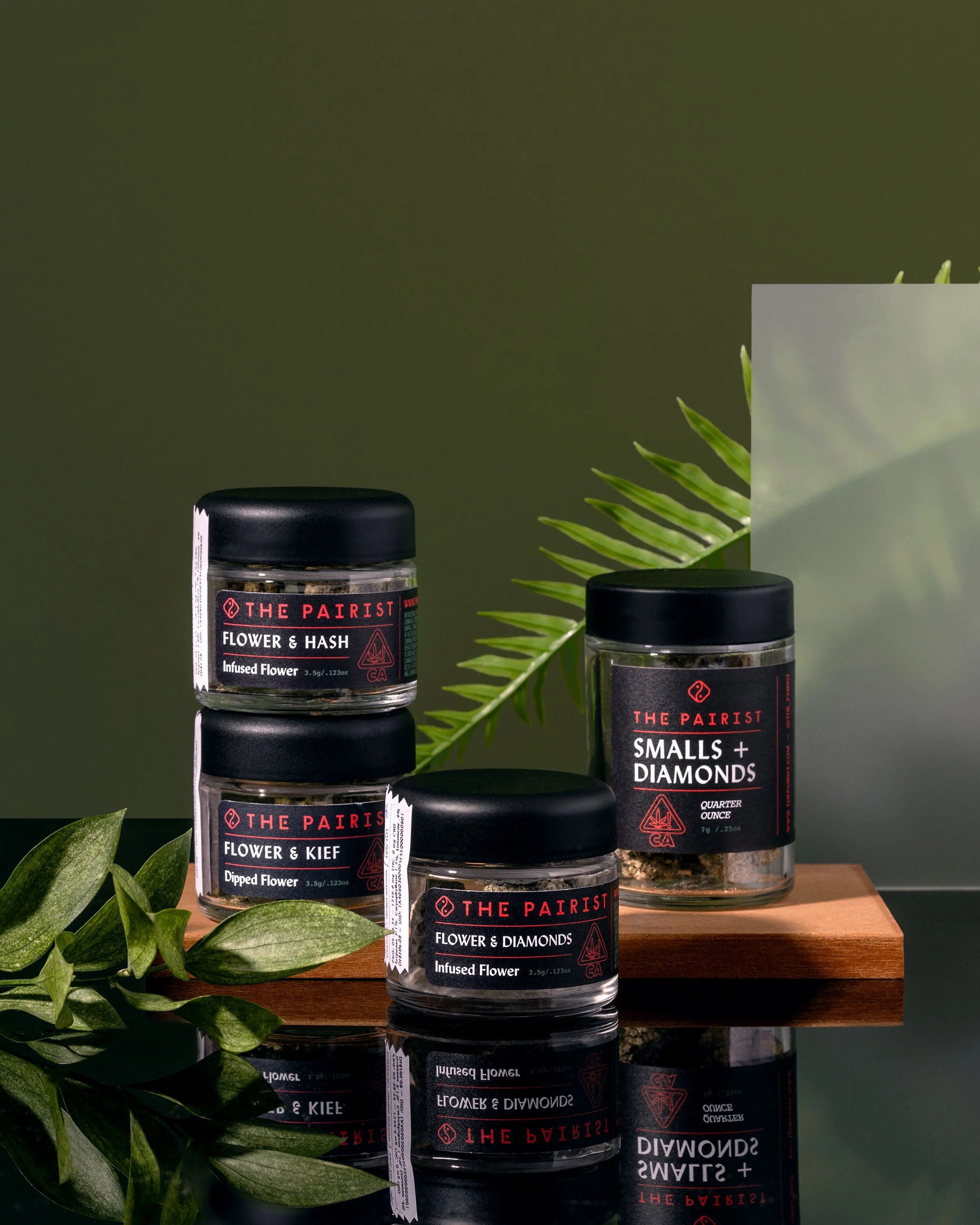 Three black-labeled jars of cannabis products, displayed on a wooden surface with green plants and a dark green background.