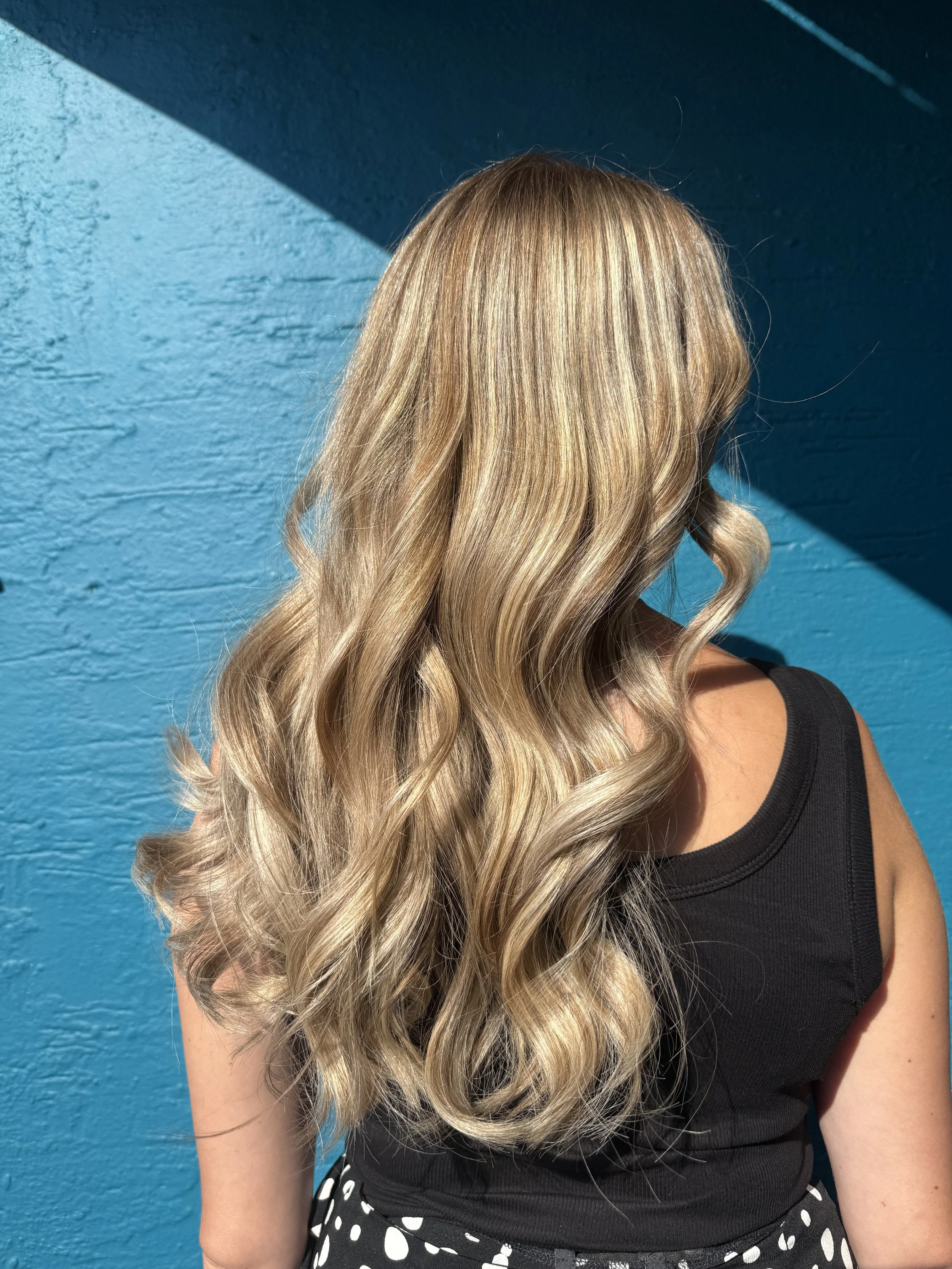 A person with long, wavy blonde hair wearing a black tank top and polka dot pants standing against a blue textured wall with a shadow.