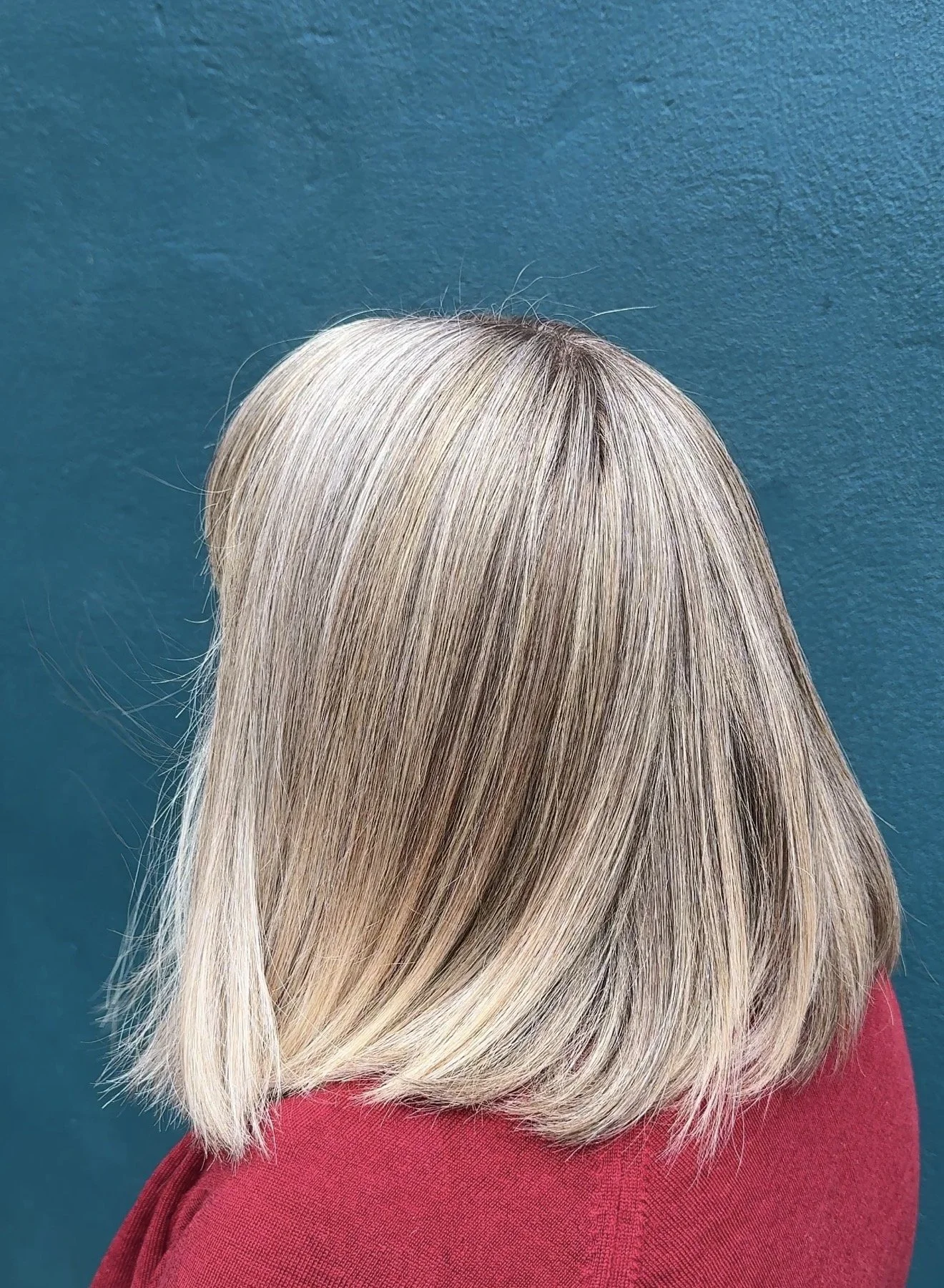 Blonde person with straight grey blended hair wearing a red top against a blue wall.
