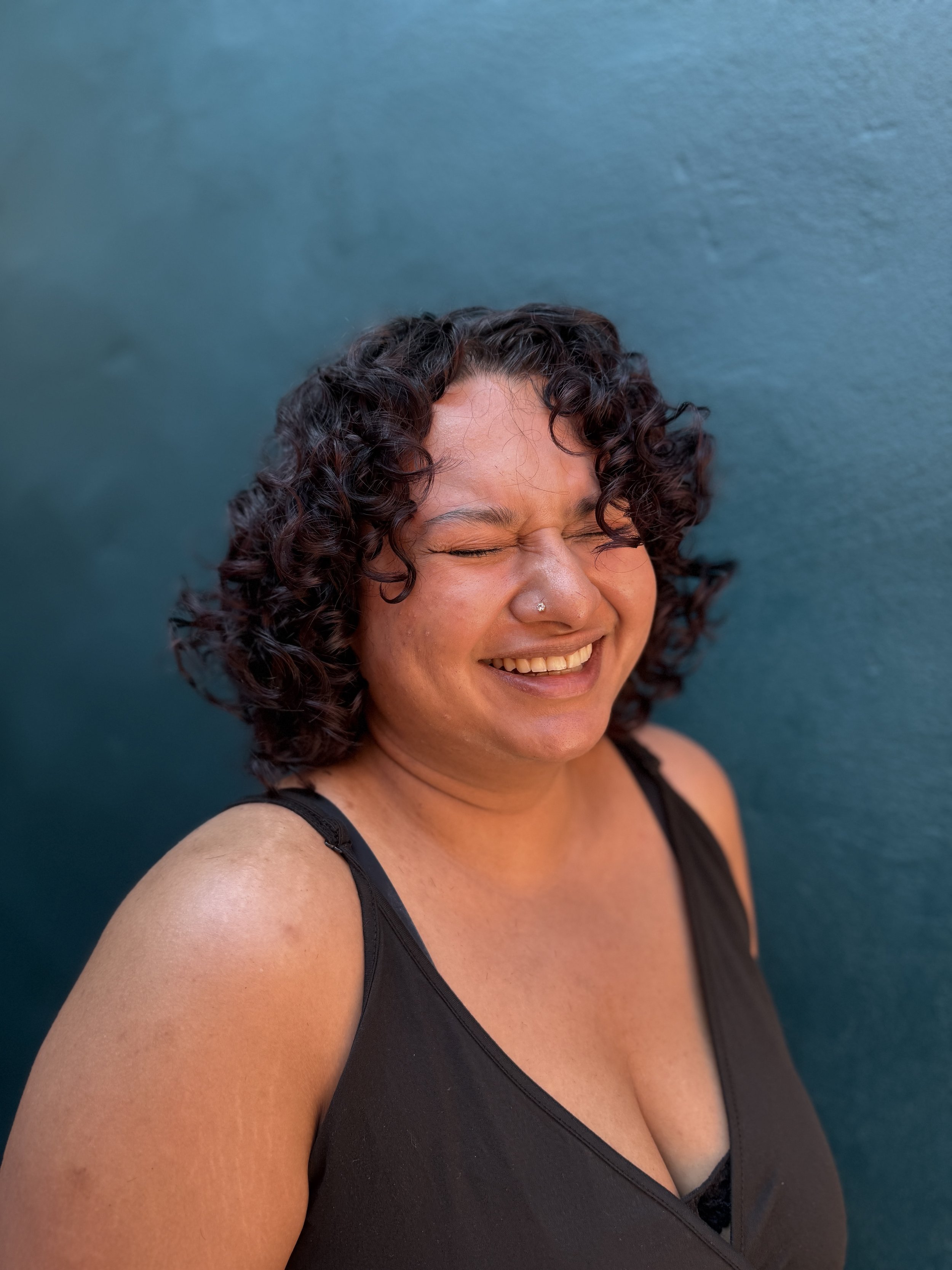 A person with curly dark hair smiling with eyes closed, wearing a black top with thin straps, standing against a teal background.