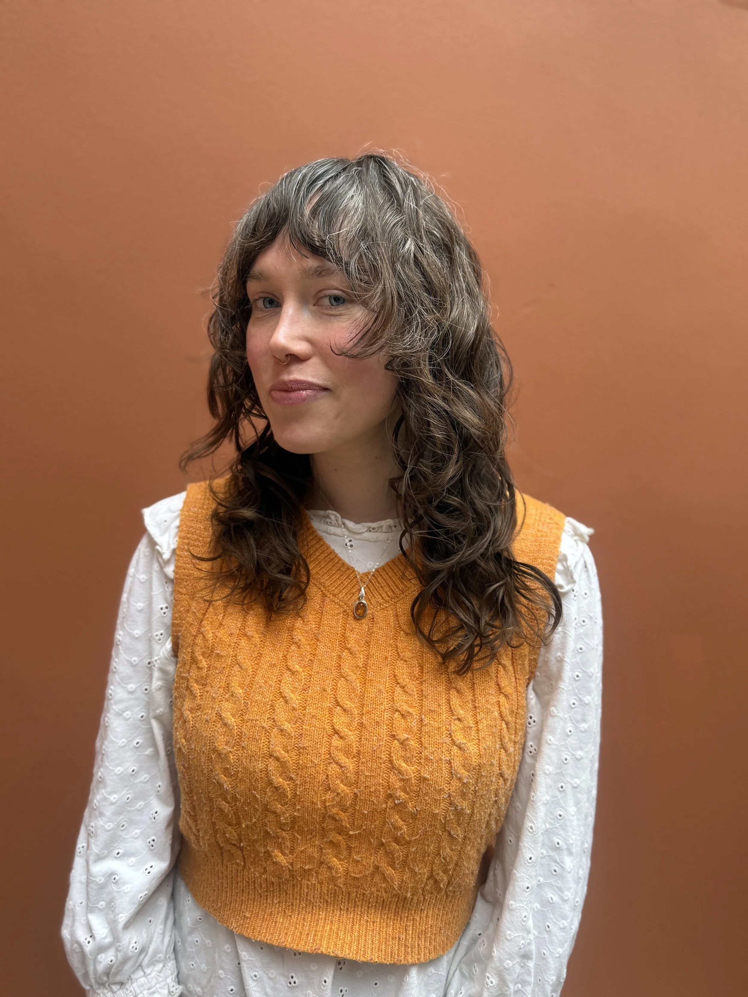 A person with curly, brown shag cut hair and blue eyes, wearing a white eyelet blouse and an orange cable-knit vest, standing against a plain brownish-orange background.