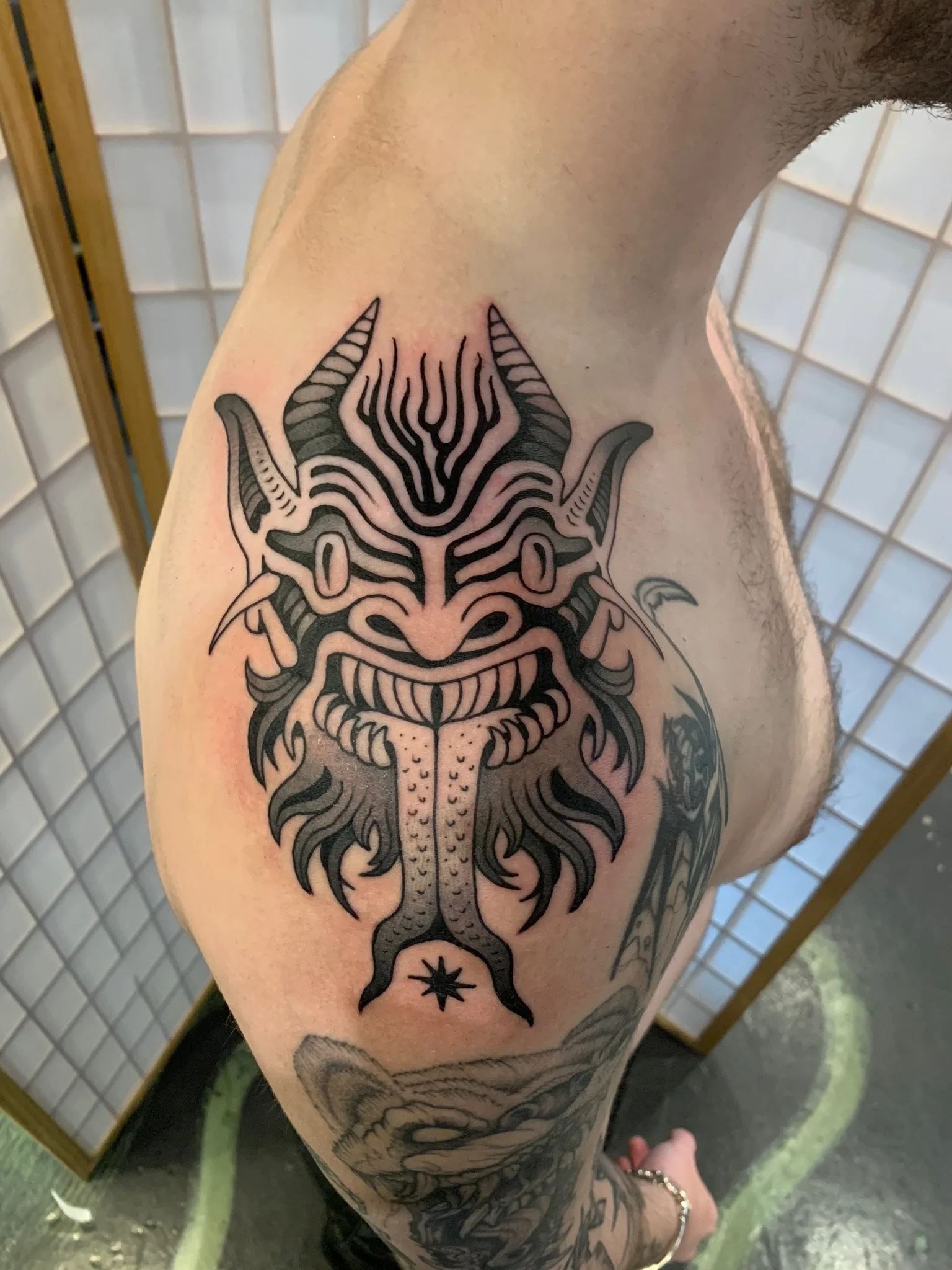 Black tribal-style tattoo of a fierce creature's face with horns, sharp teeth, and a tongue, on a person's upper arm and shoulder, made at Thanks Tattoo