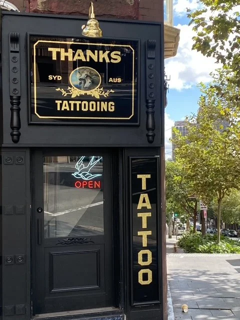 Signboard advertising tattooing services outside Thanks Tattoo shop, with a smaller neon "OPEN" sign in the door window and a gold Buddha figure on top.