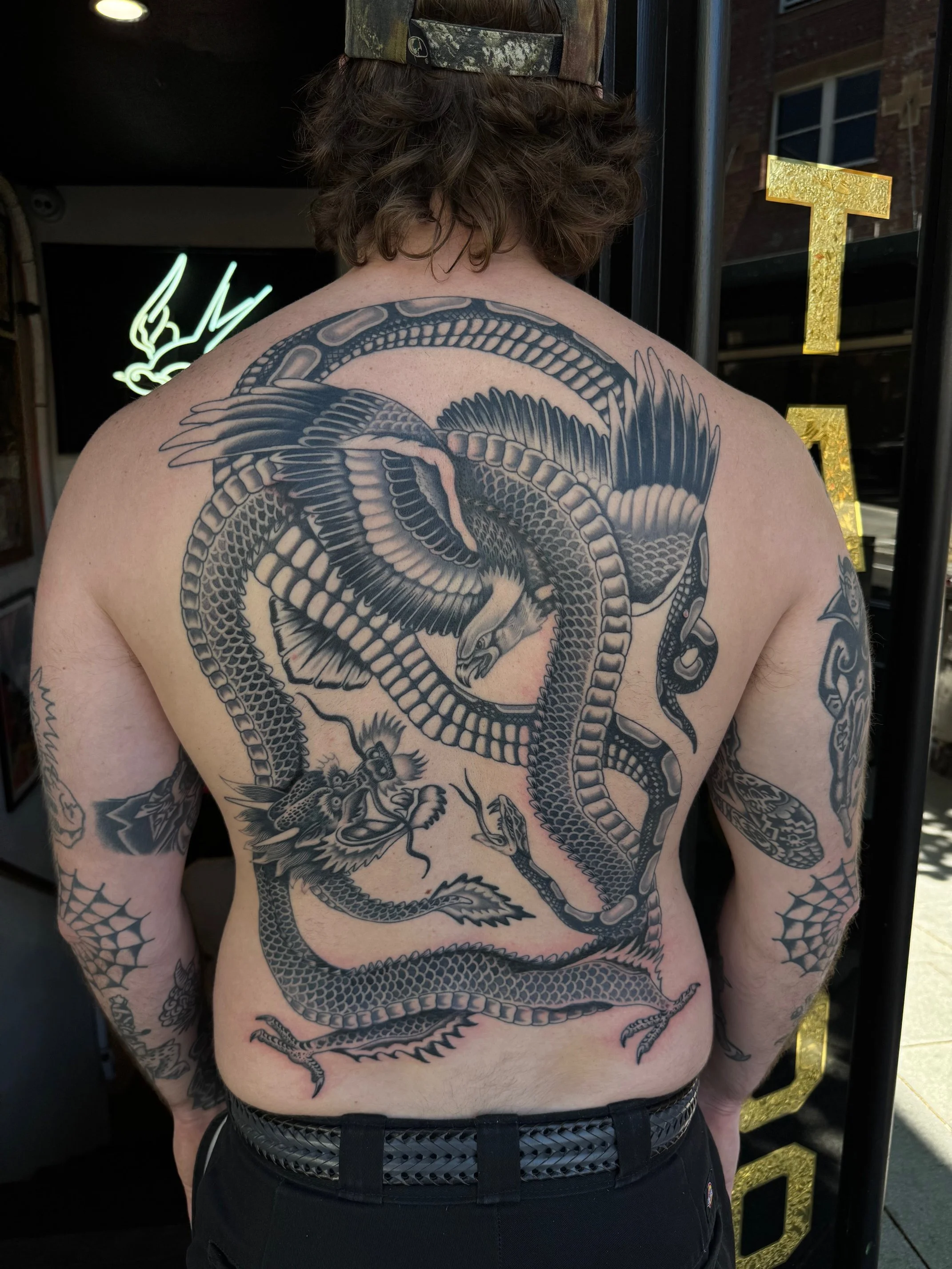 Back tattoo of a large serpent coiled around a dragon, with detailed scales and feathers, in black ink, made at Thanks Tattoo