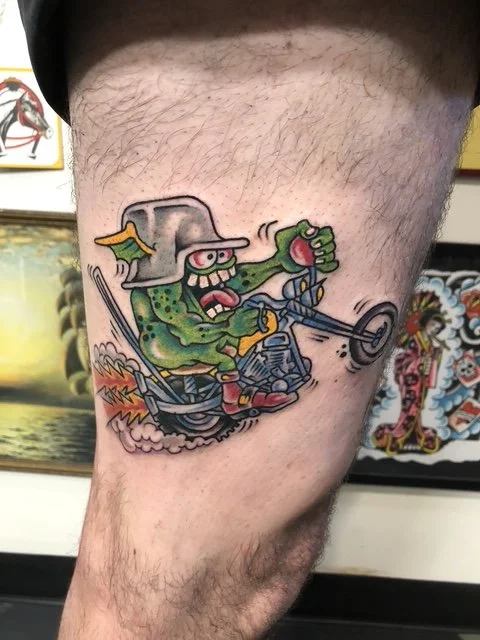 Colorful tattoo of a cartoonish witch riding a motorcycle, wearing a gray hat with a yellow feather, and making an aggressive face, made at Thanks Tattoo