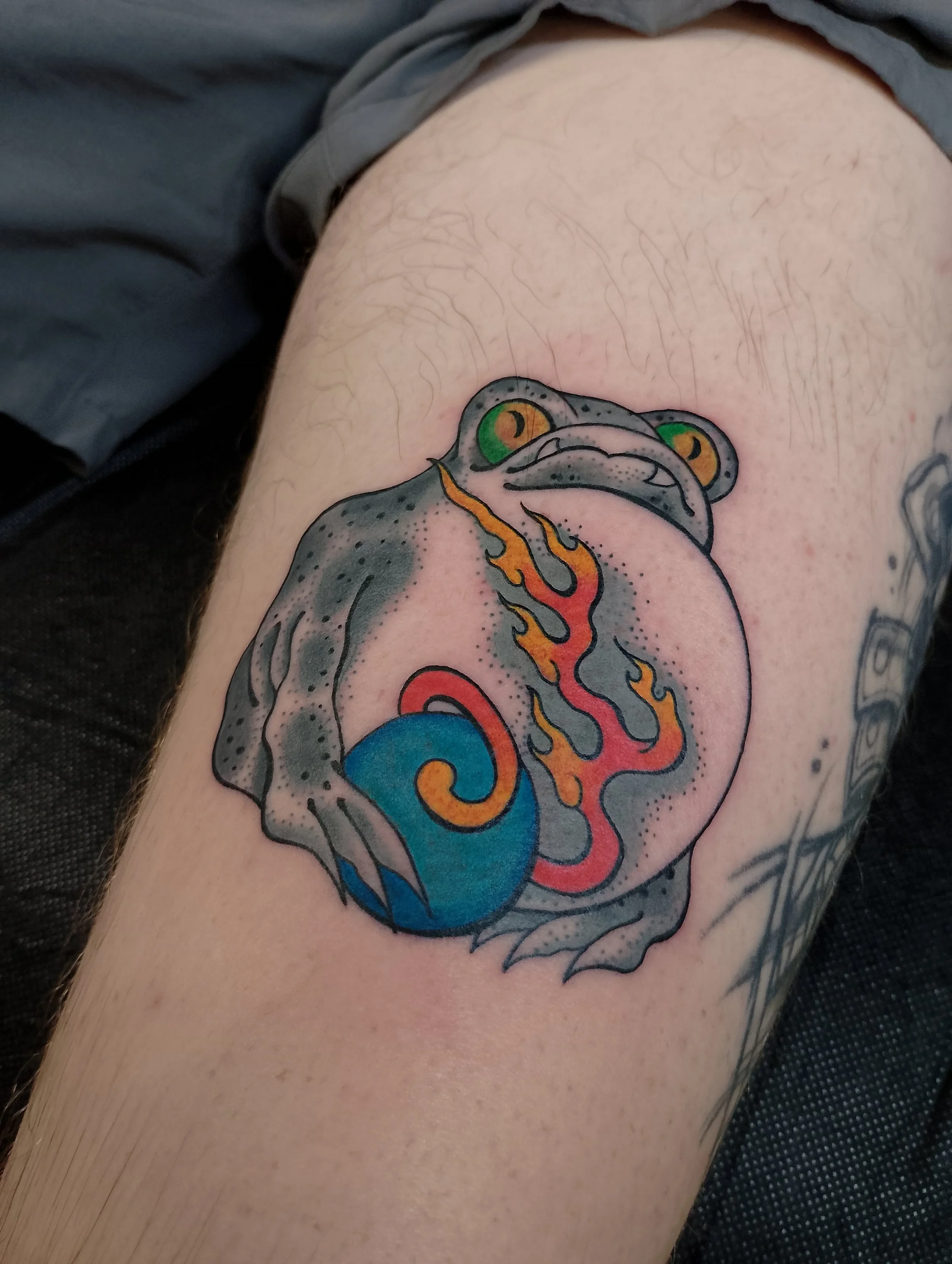 Colorful tattoo of a frog with a fire coming out of its mouth, sitting on a blue sphere, on a person's thigh, made at Thanks Tattoo