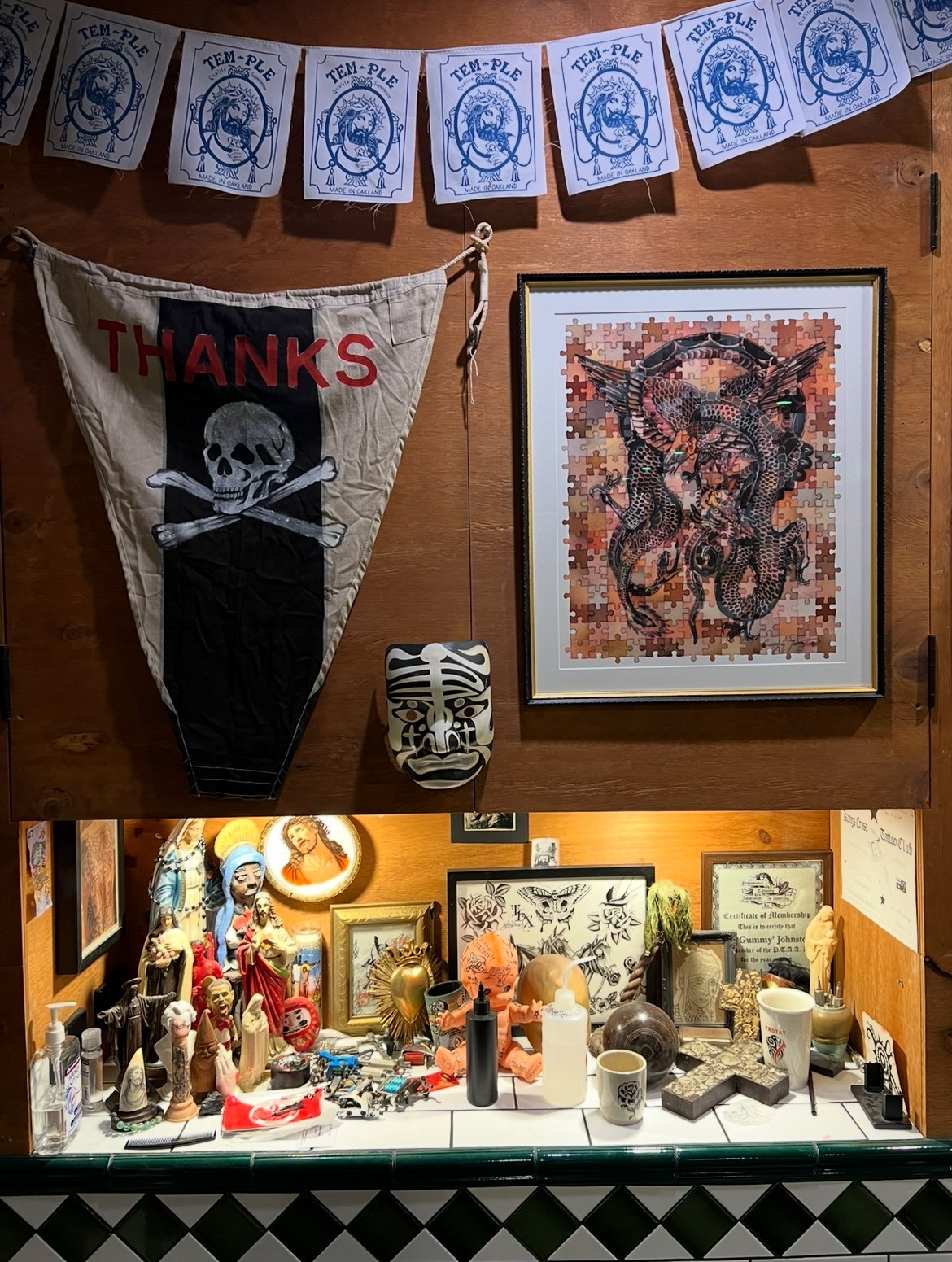 Wall with decorative items including a framed painting of snakes, a 'Thanks' banner with a skull and crossbones, a carved mask, and various religious statues and figurines. The bottom shelf has small statues, candles, pictures, and decorative objects