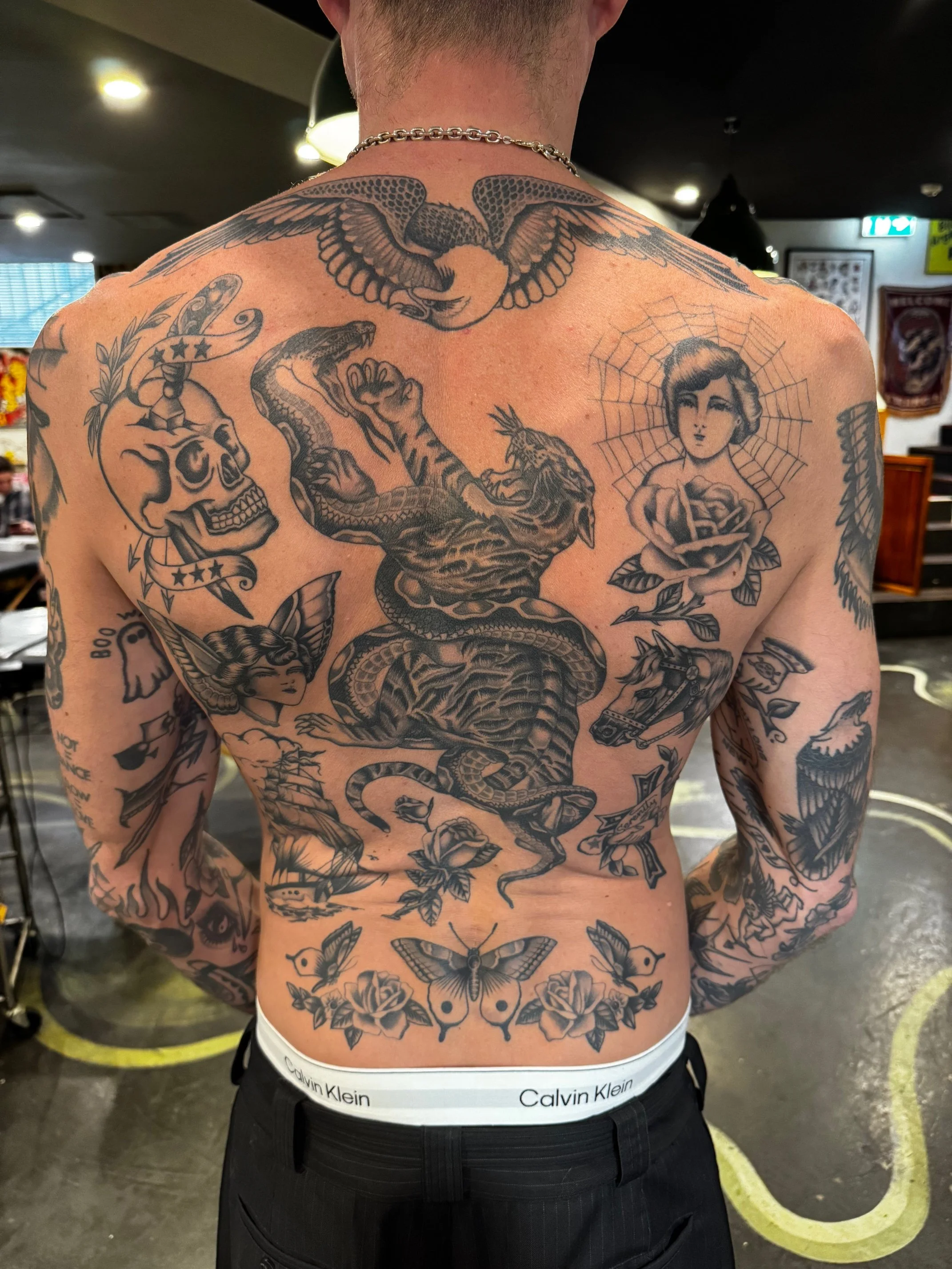The tattoos include a flying bird, a skull with a top hat, a woman with a spider web halo, a snake wrapped around a muscular creature, roses, butterflies, a gun, and various other designs, made at Thanks Tattoo