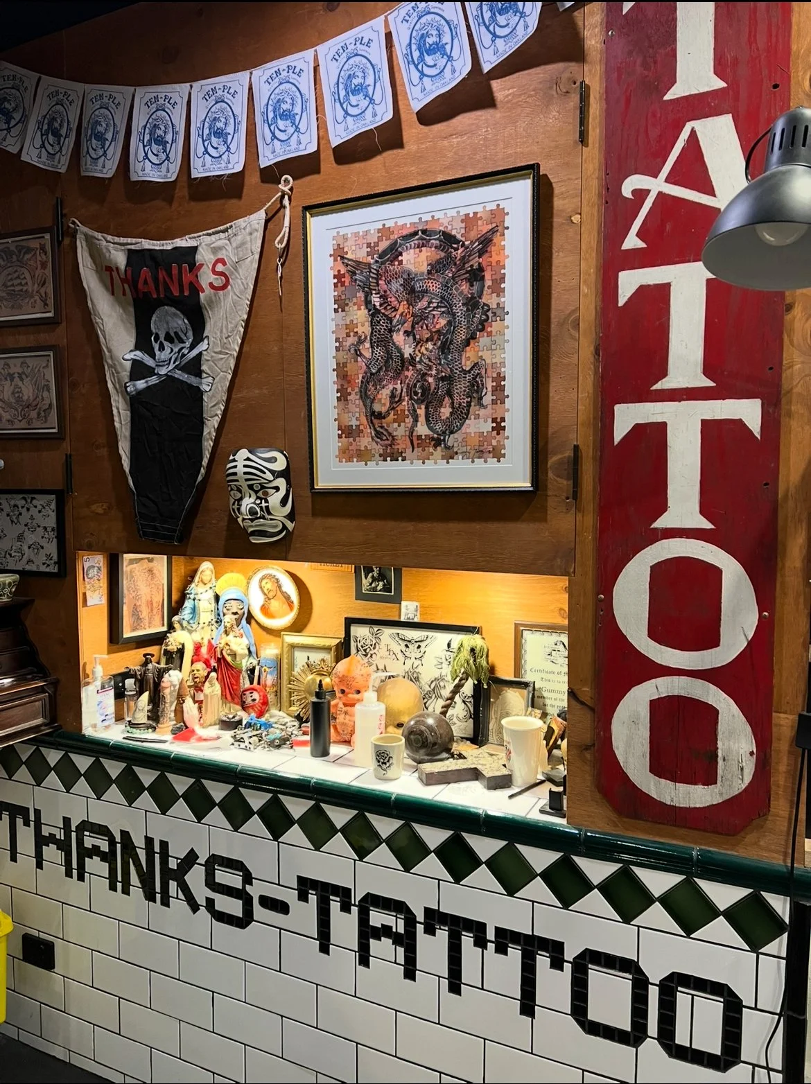 decorated interior with various artworks and objects. There is a framed puzzle with a dragon and four wings on the wall, a fabric banner with a skull and crossbones, and a string of flags with a blue crest pattern. Below, a counter with religious sta