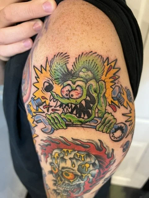 Colourful tattoo of a cartoonish, green, devil-like creature with a mischievous expression, sharp teeth, and horns, surrounded by various objects and flames on a person's upper arm, made at Thanks Tattoo