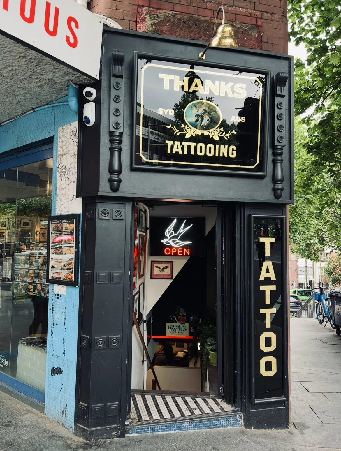 Exterior of a tattoo studio with a black entrance, a neon 'OPEN' sign, and signs indicating tattooing services, including a large sign reading 'Thanks Tattooing' with a unicorn logo, and a vertical sign spelling 'TATTOO' on the right side of the entr