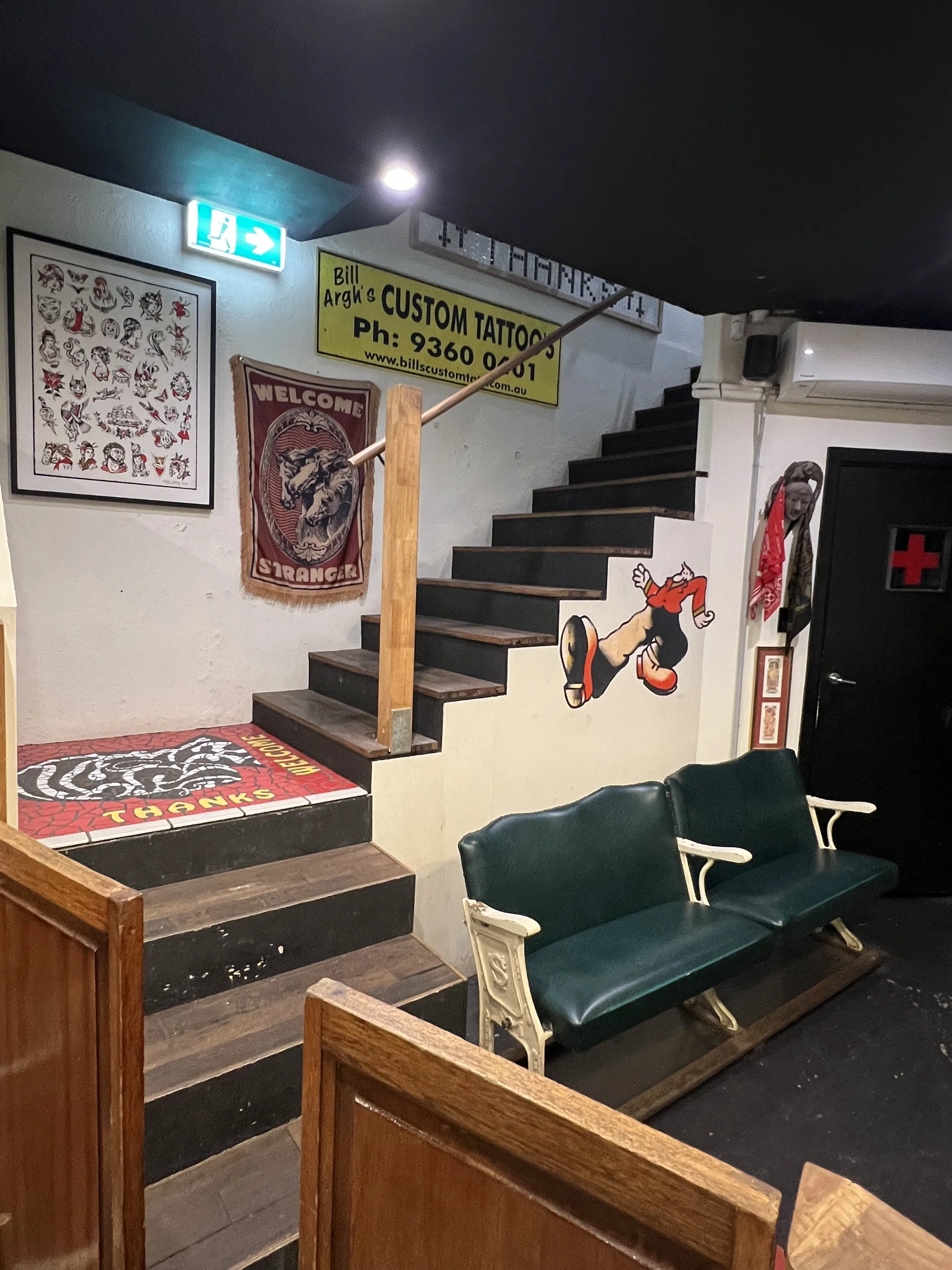 Interior of a Thanks tattoo shop with staircase, tattoo artist signs, wall art including a wolf banner, cartoon character decal, vintage statue, and seating area with green bench.